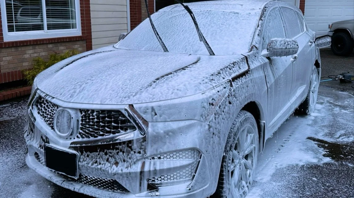 White SUV covered in soap suds during a car wash, with windshield wipers raised and soap dripping on the ground.
