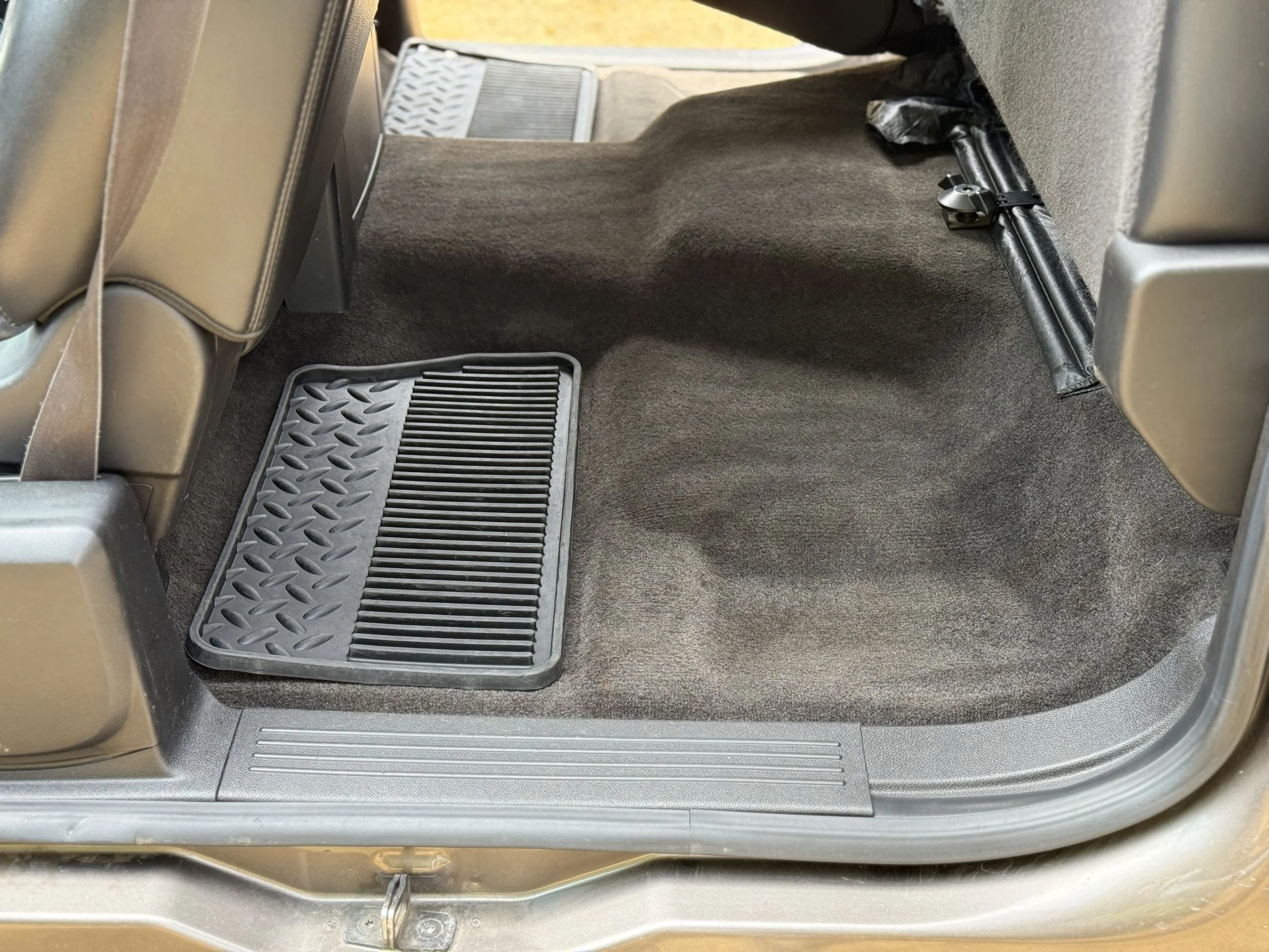 Backseat area of a car showing brown carpeted flooring, a floor mat, and part of the back of the front seats with a plastic cover on the sides.