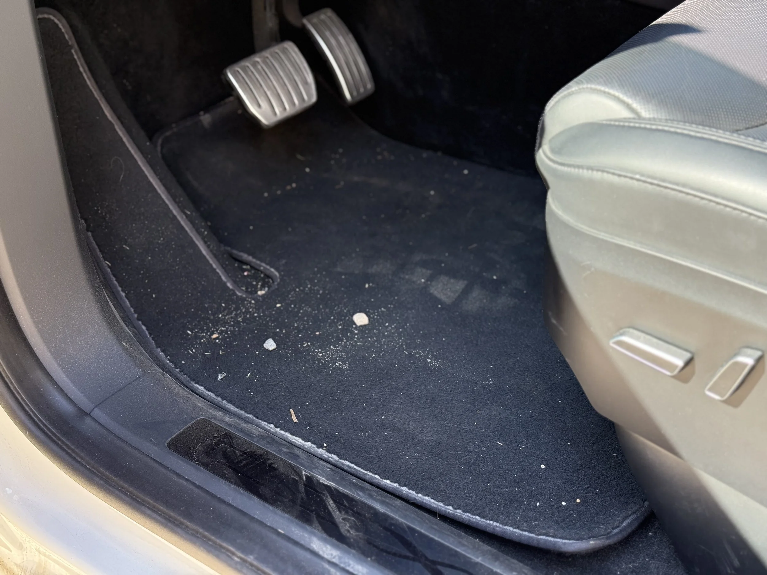 Car interior showing the driver's side footwell with pedals, carpet, and a part of the seat. The carpet has small debris and dust.