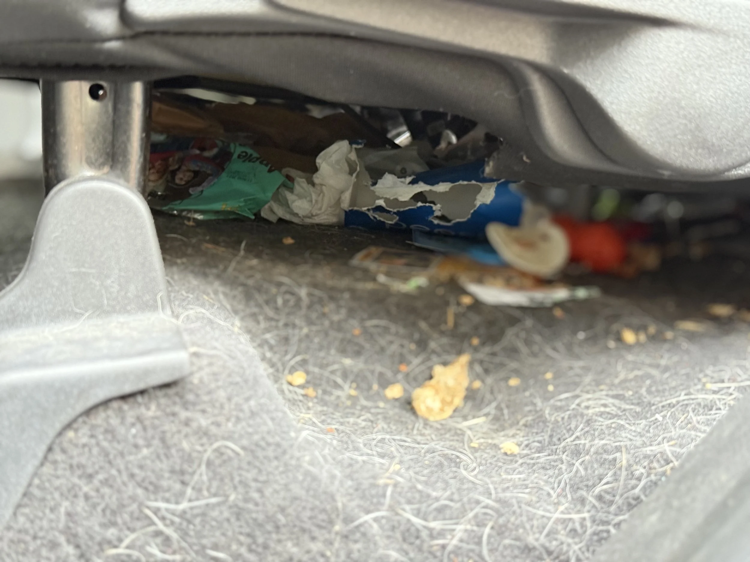 Car interior showing trash and debris under the seat, including paper, wrappers, food crumbs, and dust.