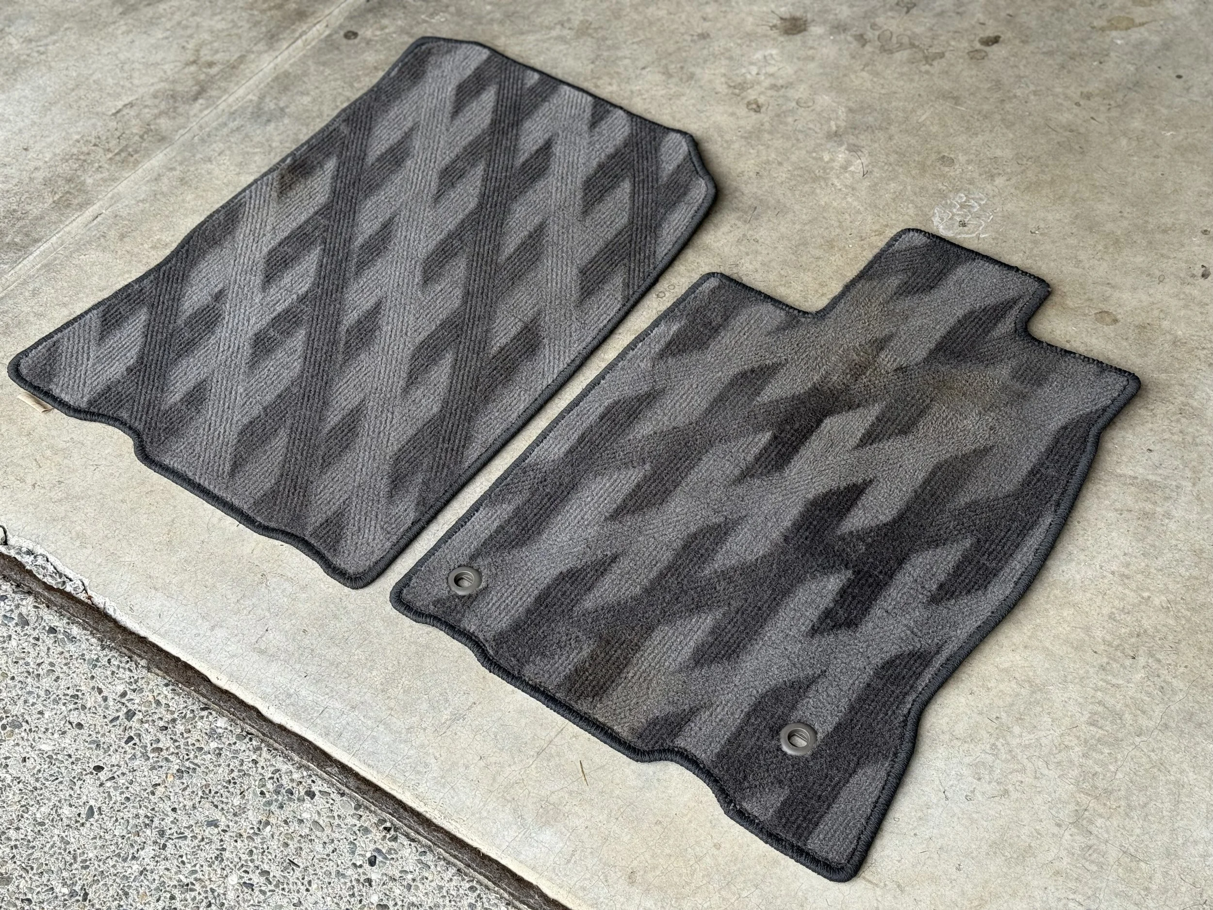 Two gray and black vehicle floor mats with a geometric pattern, placed on a concrete floor.