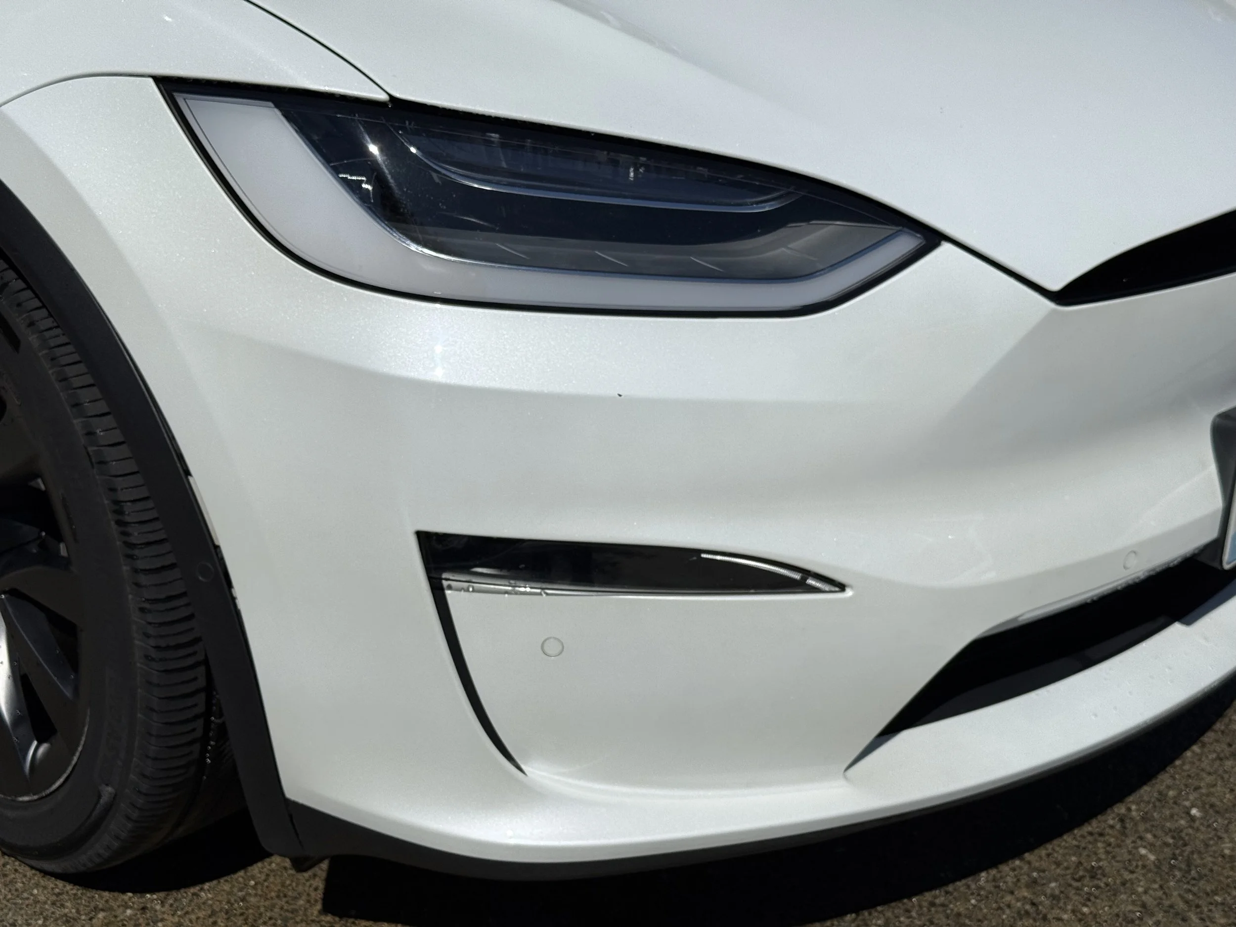 Close-up of the front left side of a white Tesla car, showing the headlight and tire.