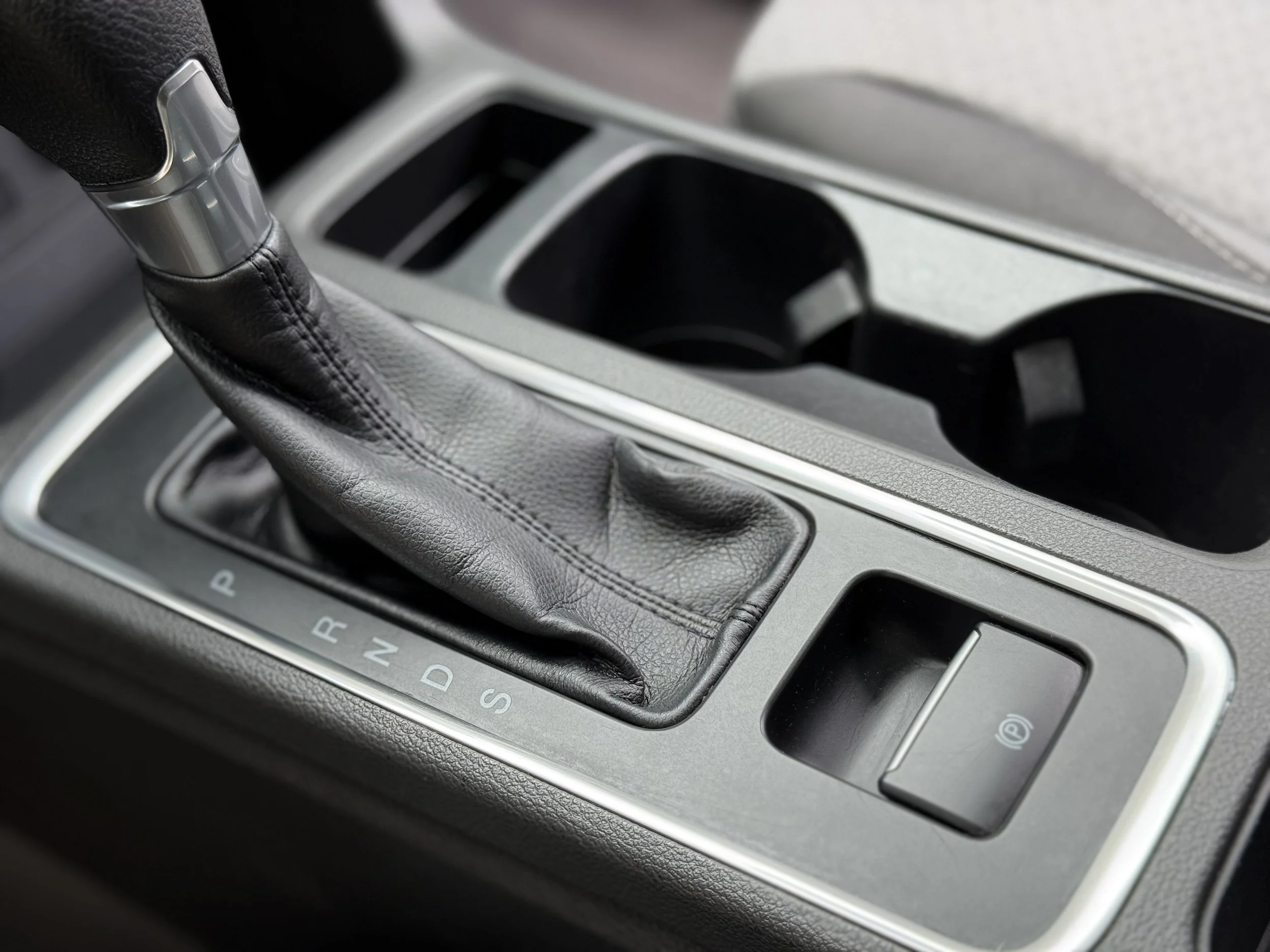Close-up of a car's gear shift lever wrapped in black leather, situated on the center console next to a parking brake button.