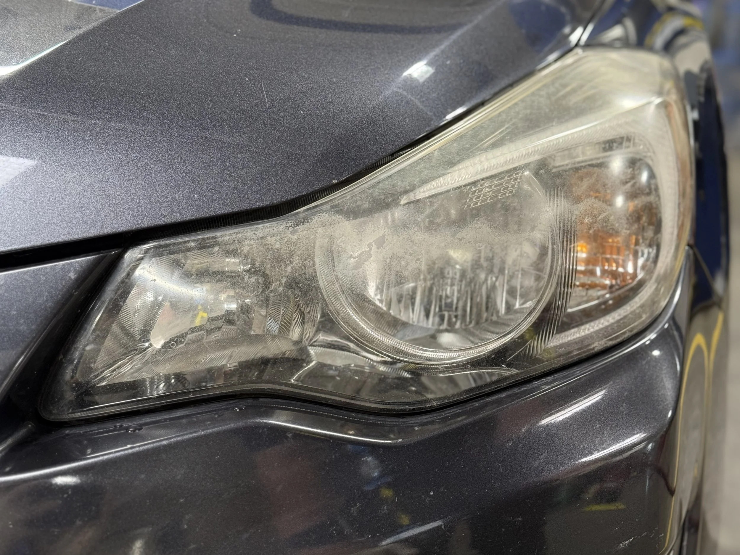 Close-up of a car's headlight with a dusty, slightly foggy lens, part of the black car body, and a reflection on the surface.