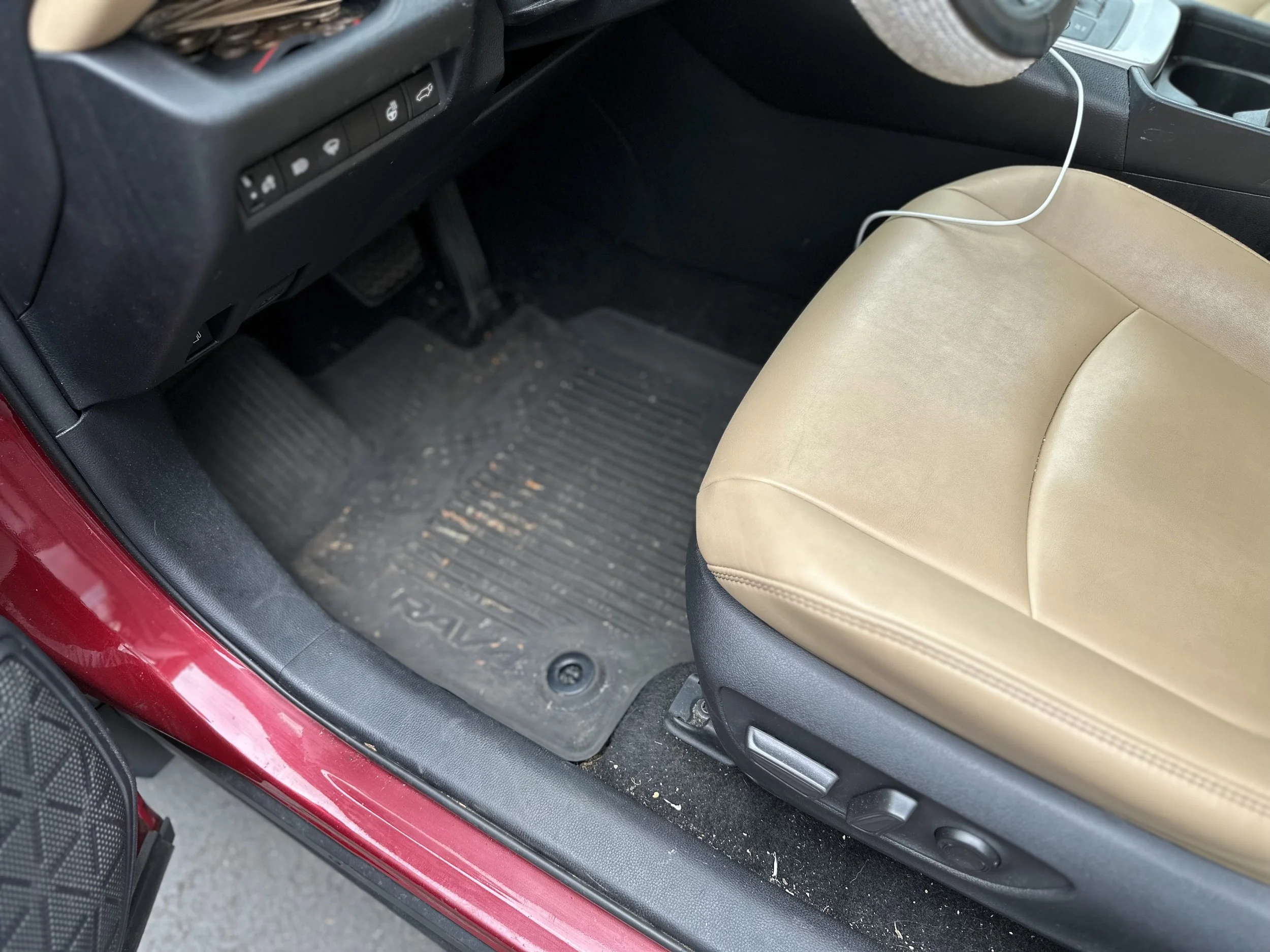 The front driver side of a car showing a black rubber floor mat inside, a beige leather seat, and part of the dashboard with buttons.