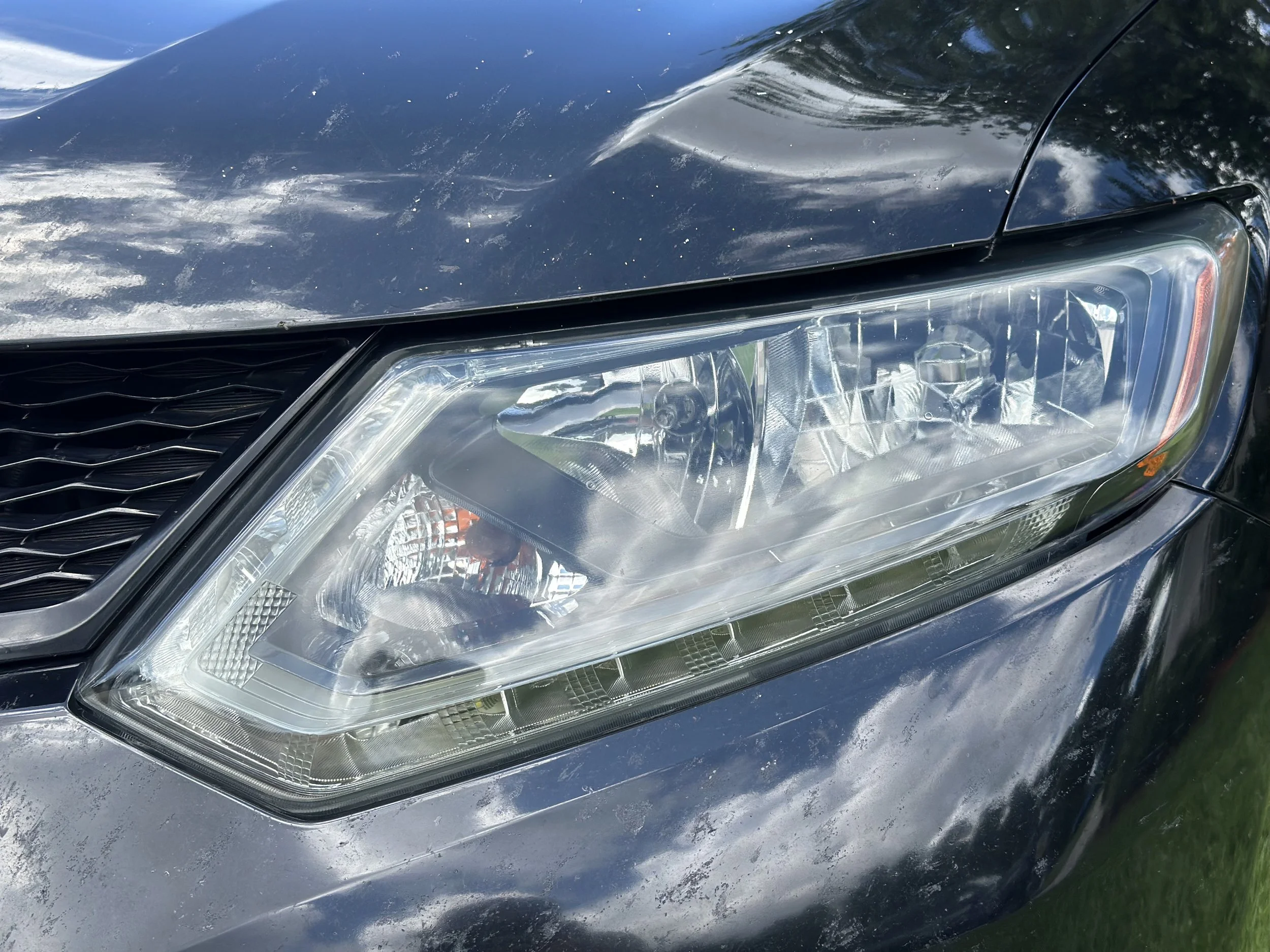Close-up of a black car's headlight and front grille with reflections of clouds and sky.