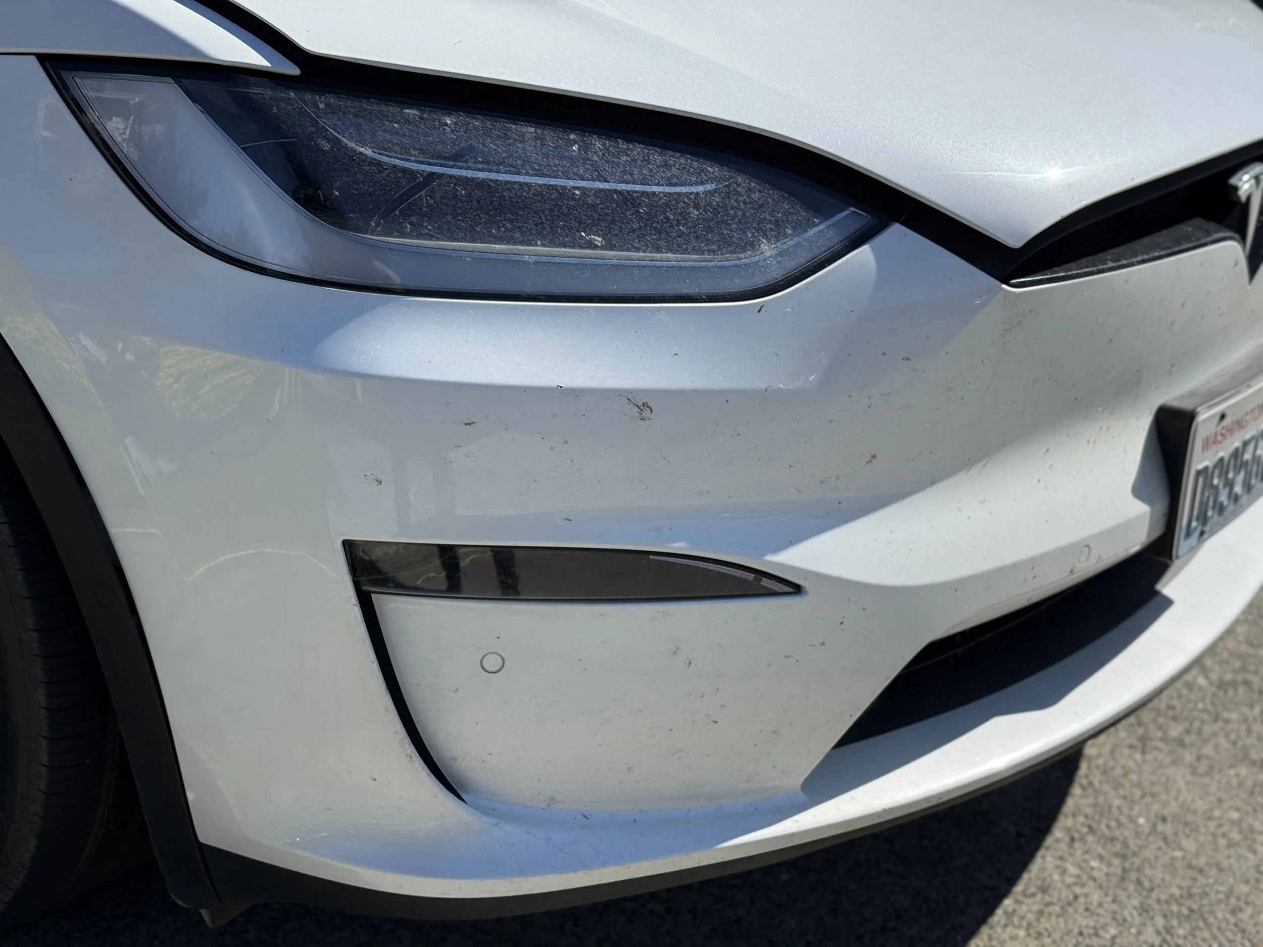 Close-up of the front left side of a white luxury car, showing the headlight, part of the grill, and the front bumper with some dirt and small scratches.