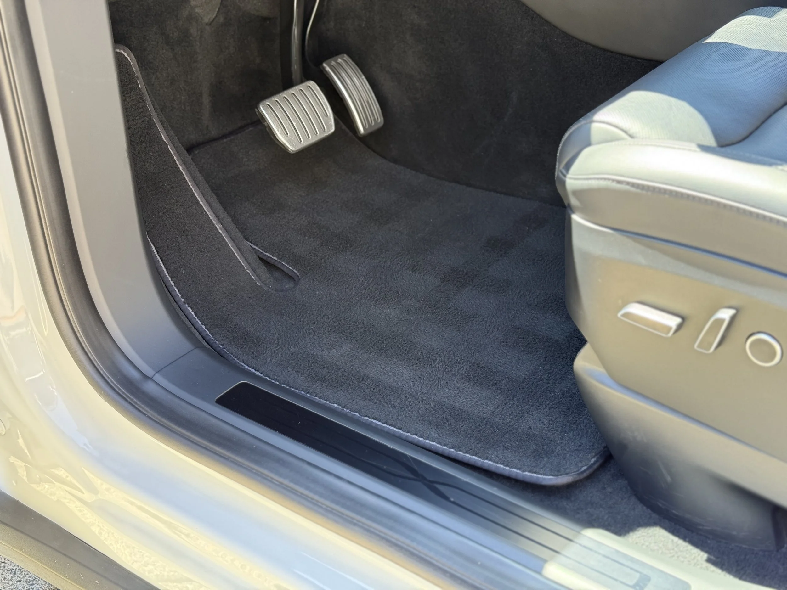 Close-up of a car interior showing driver’s side floor area with accelerator and brake pedals, gray side of a seat with control buttons, and a black floor mat.