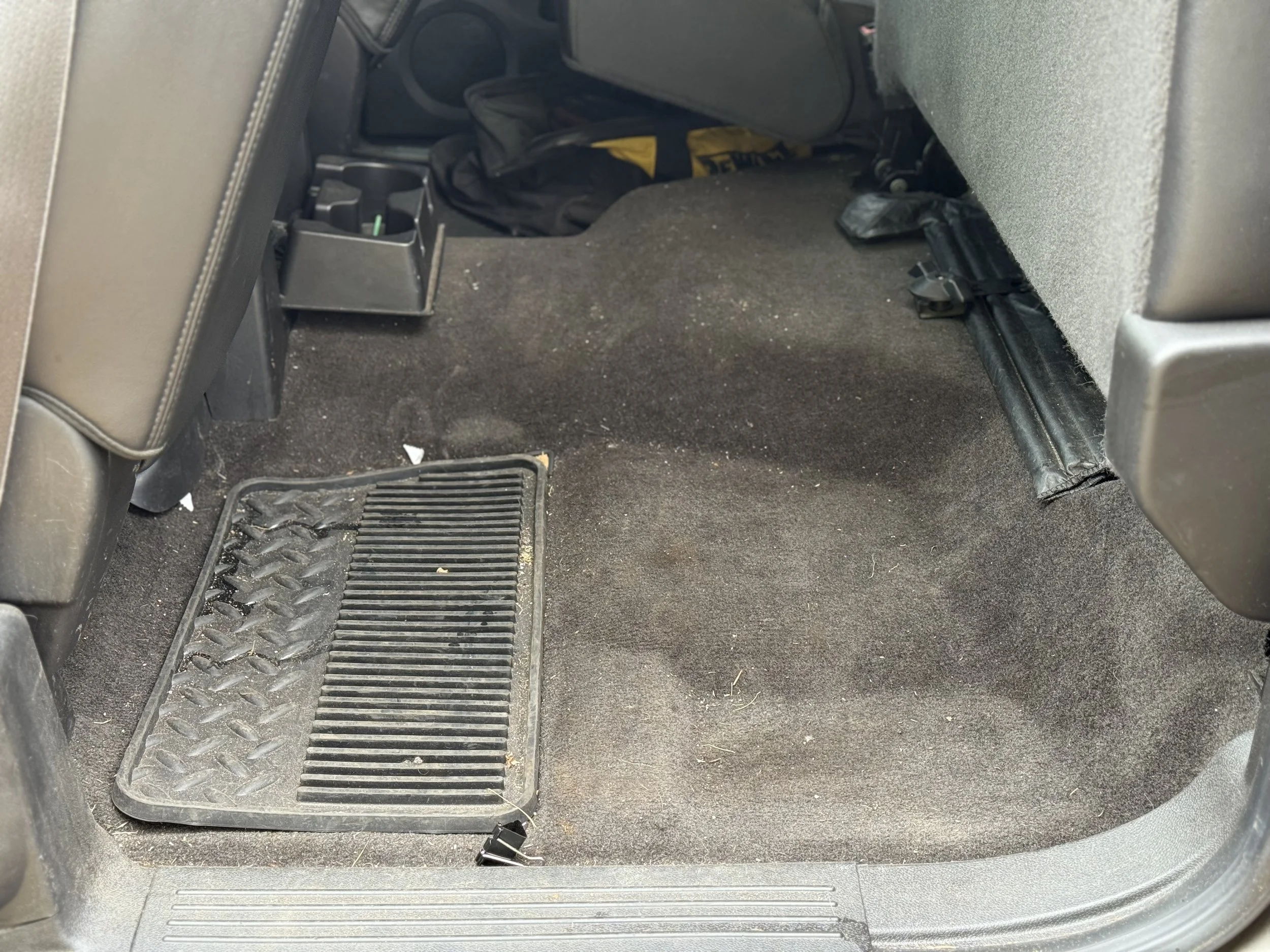 Car interior showing rear floor with black carpet, a rubber floor mat, some dirt, small debris, and part of the back of the front passenger seat.
