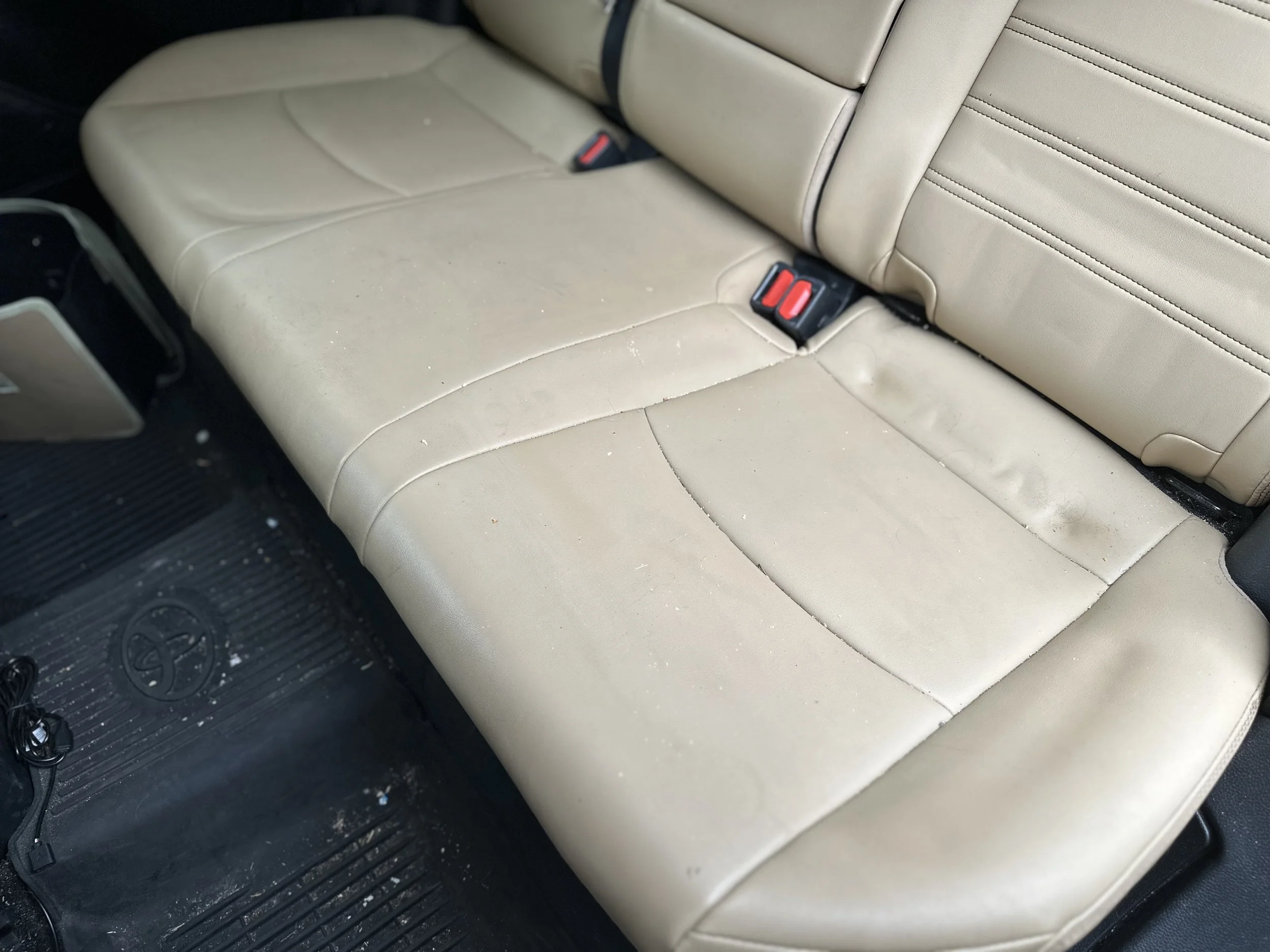 The back seat of a car with beige leather upholstery that is stained and dirty, with a black rubber floor mat below.
