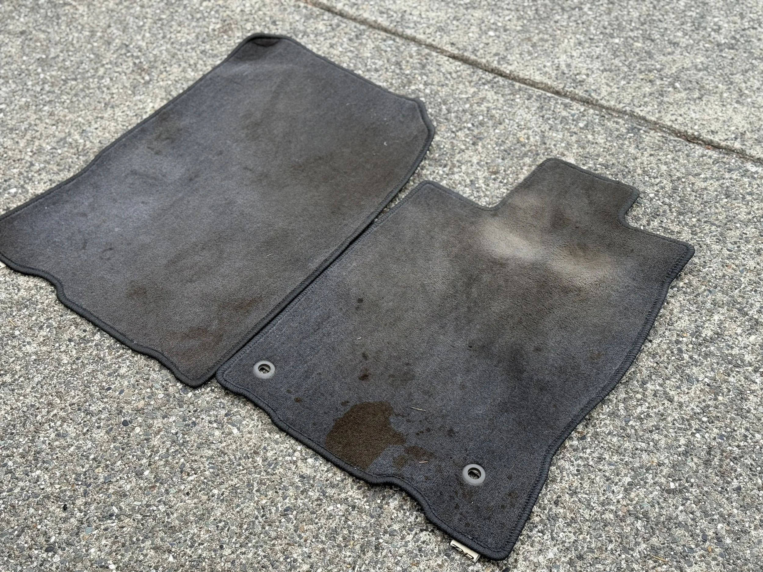 Two dirty black car floor mats on a concrete sidewalk.