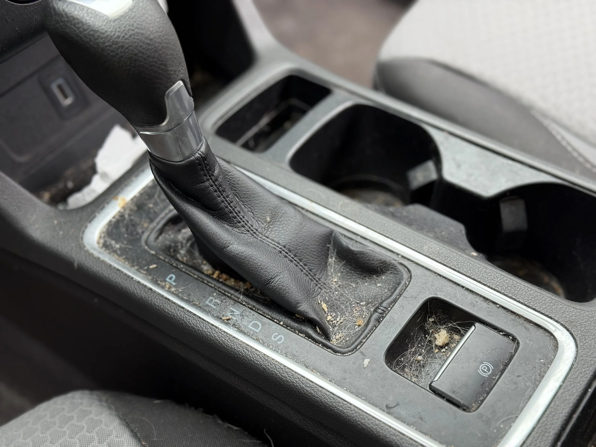 Dirty car gear shift area with debris and dust on the console and around the parking brake button.