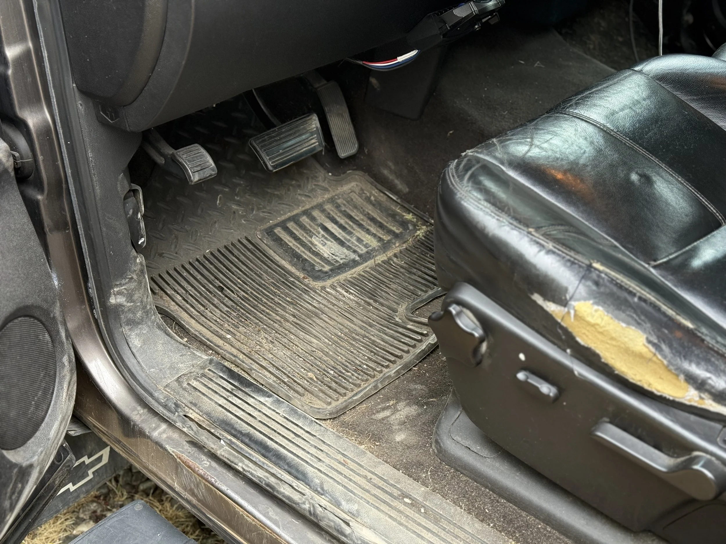 The interior of a vehicle showing the driver's side floor, with worn and dirty rubber floor mats, gas and brake pedals, and a black seat with torn upholstery.