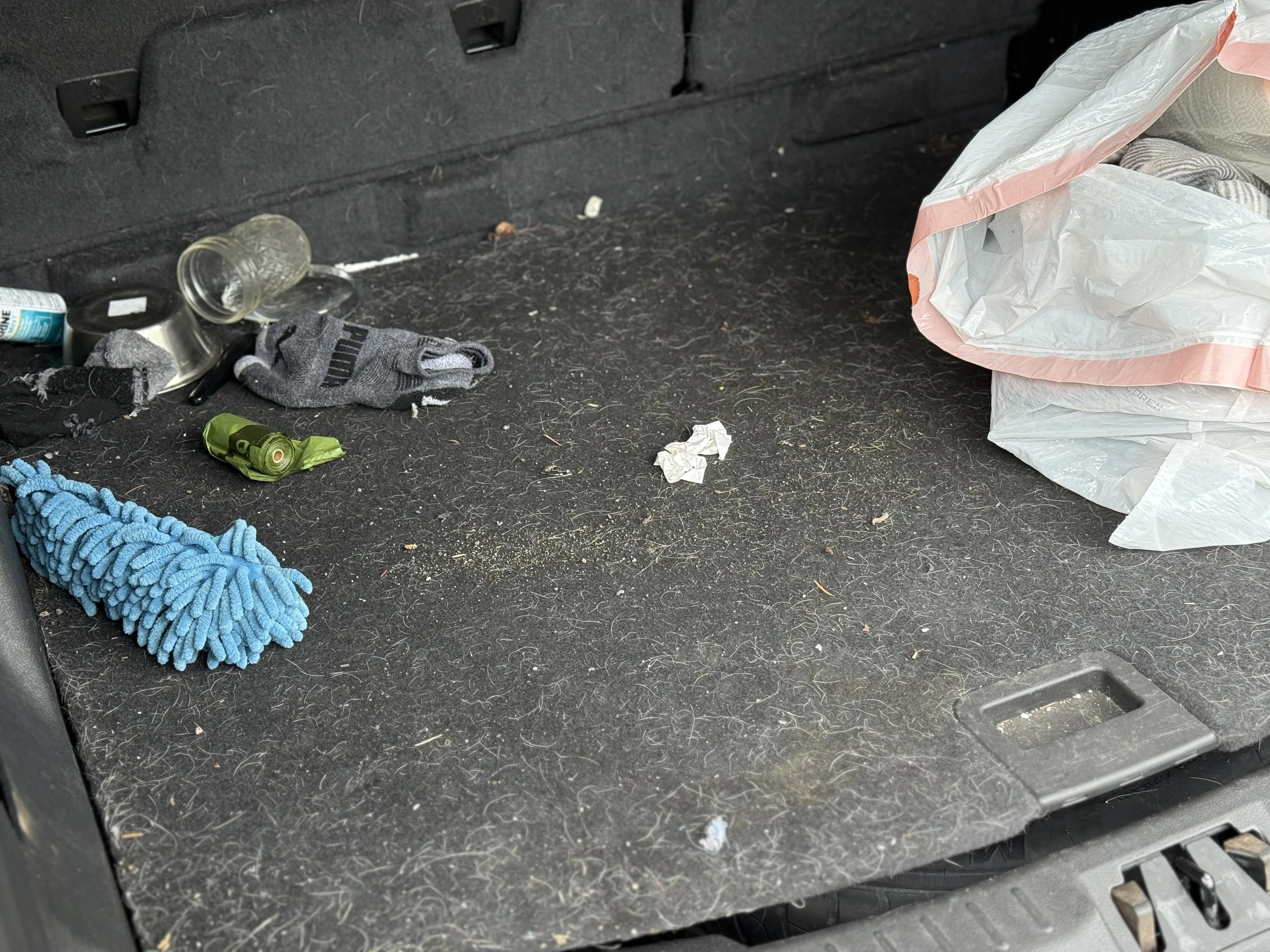A dirty car interior cargo area with trash and random items, including a blue microfiber cloth, a gray glove, a small glass jar, a crumpled tissue, and a plastic bag.