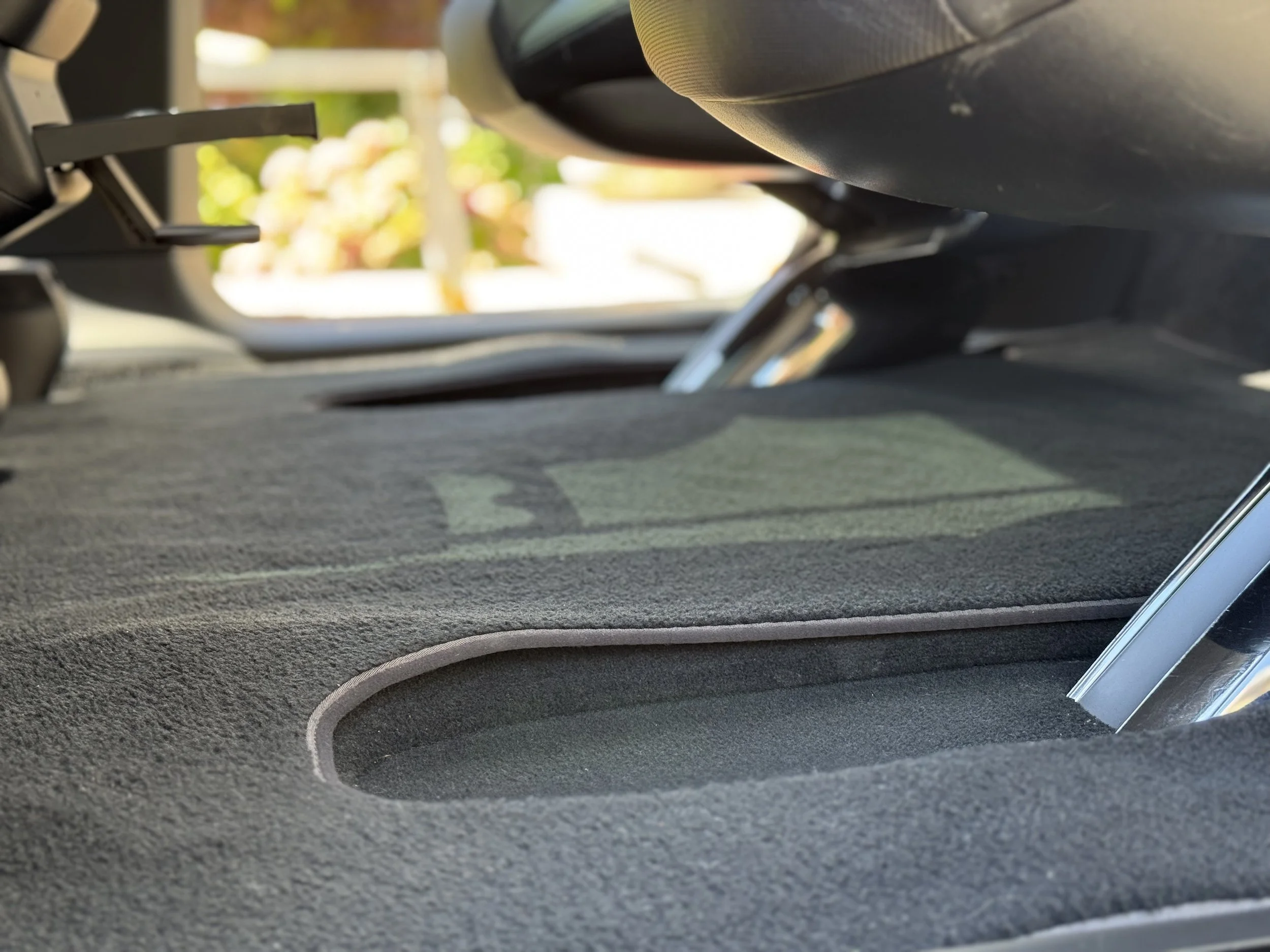View of the interior of a vehicle from the backseat, focusing on the floor and under the front seats, with a blurred outdoor background visible through the open rear door.