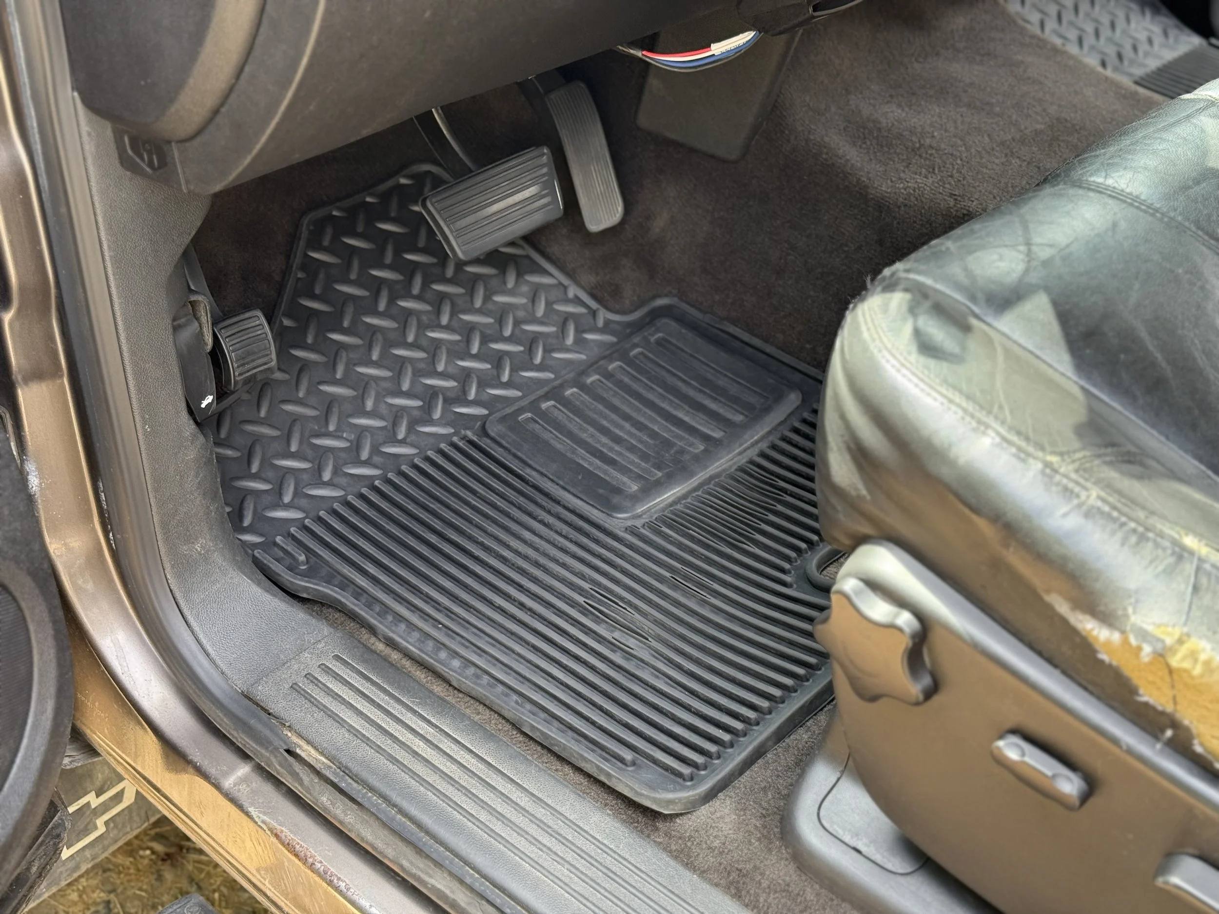 The interior of a vehicle showing the driver's side floor with three pedals, a rubber floor mat, and part of a worn leather seat.