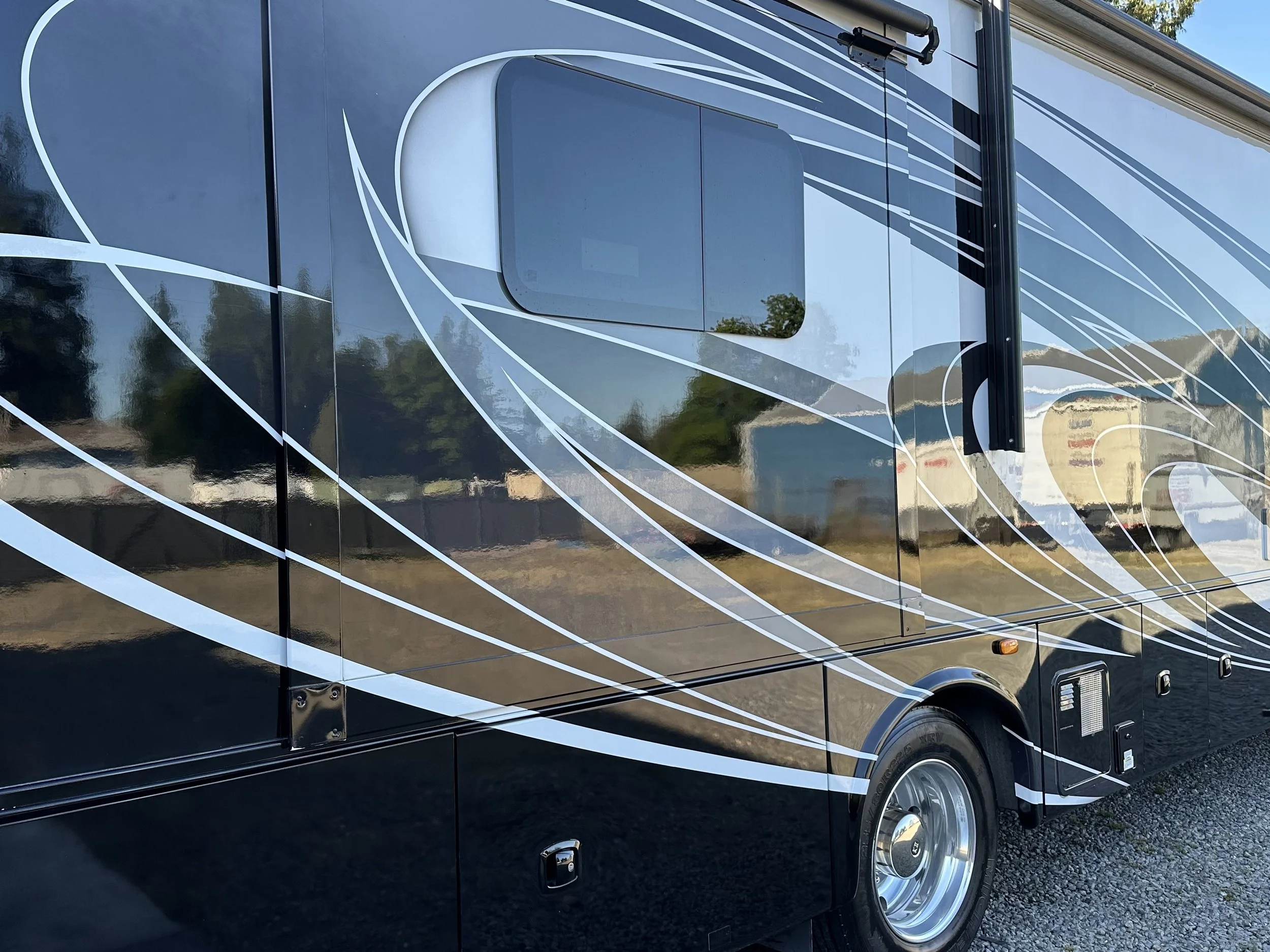 The image shows a black RV or motorhome with decorative white and silver swirl patterns on its side. It has a window and a side mirror visible, with a reflection of the surrounding landscape on its shiny surface.