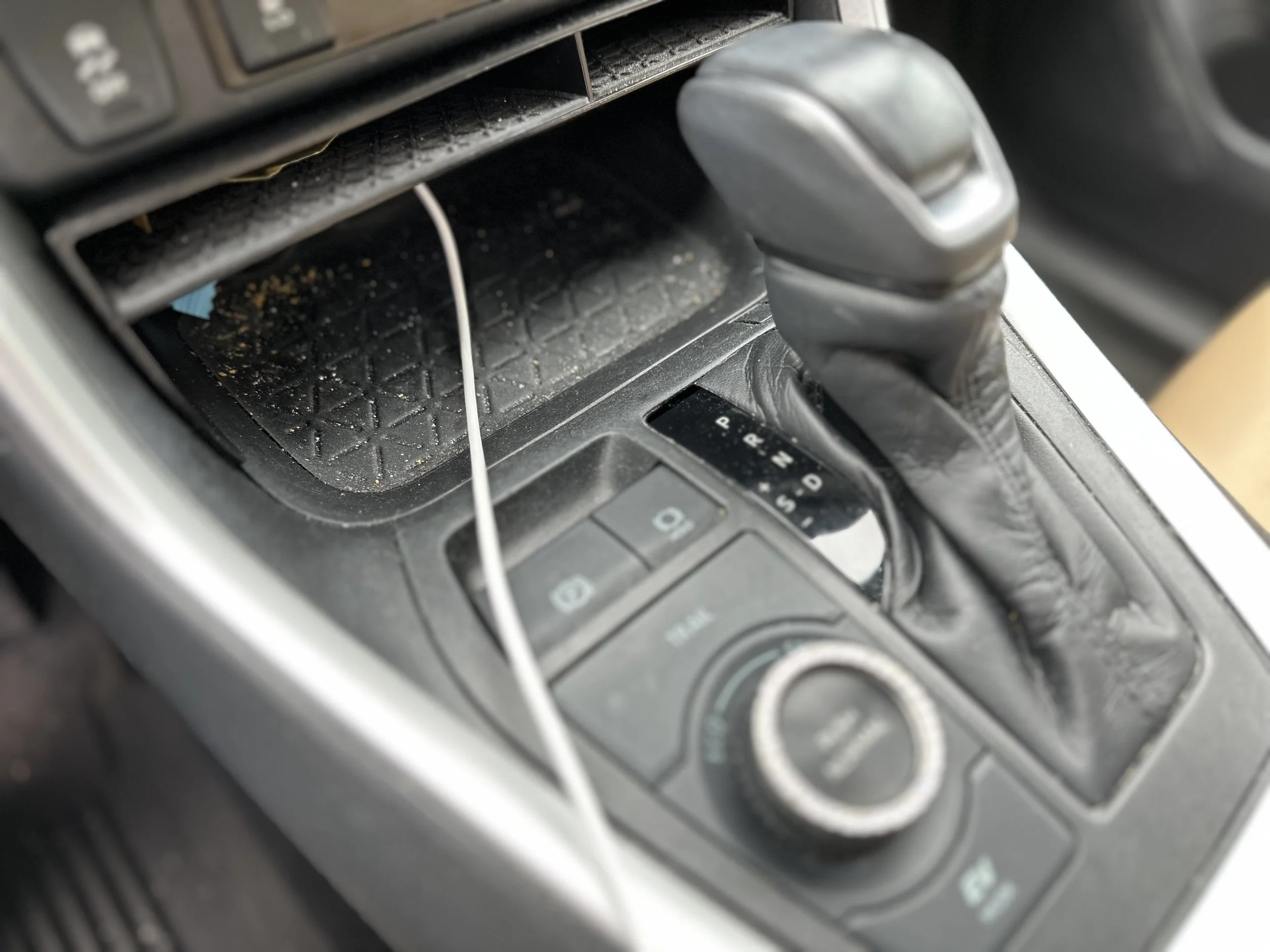 Center console of a car interior with gear shift, control buttons, and an empty storage compartment with dirt and a loose cable inside.