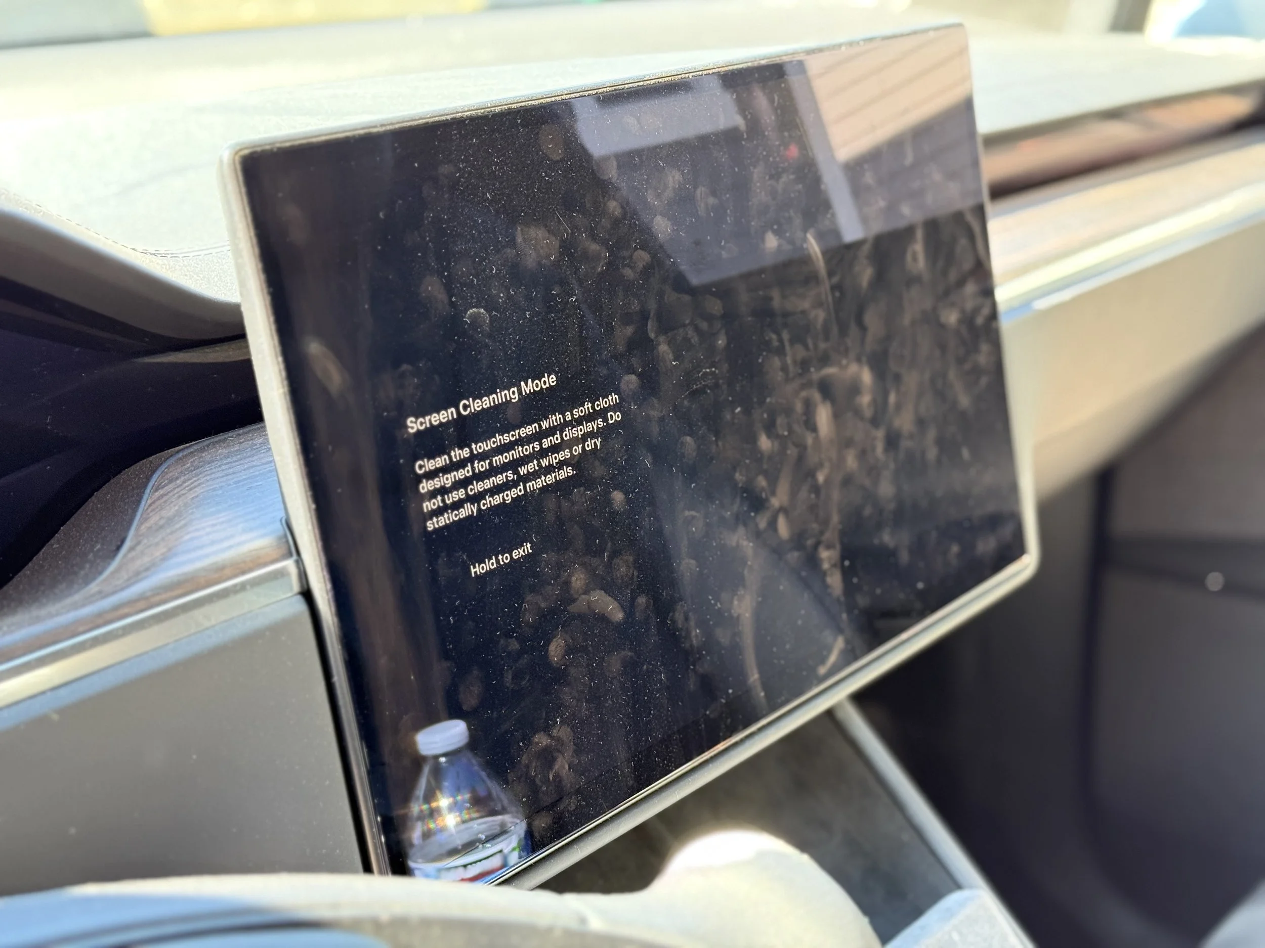 Close-up of a touchscreen in a car displaying the 'Screen Cleaning Mode' instructions, with a dusty and smudged screen, a water bottle reflection, and the car's interior and dashboard visible.