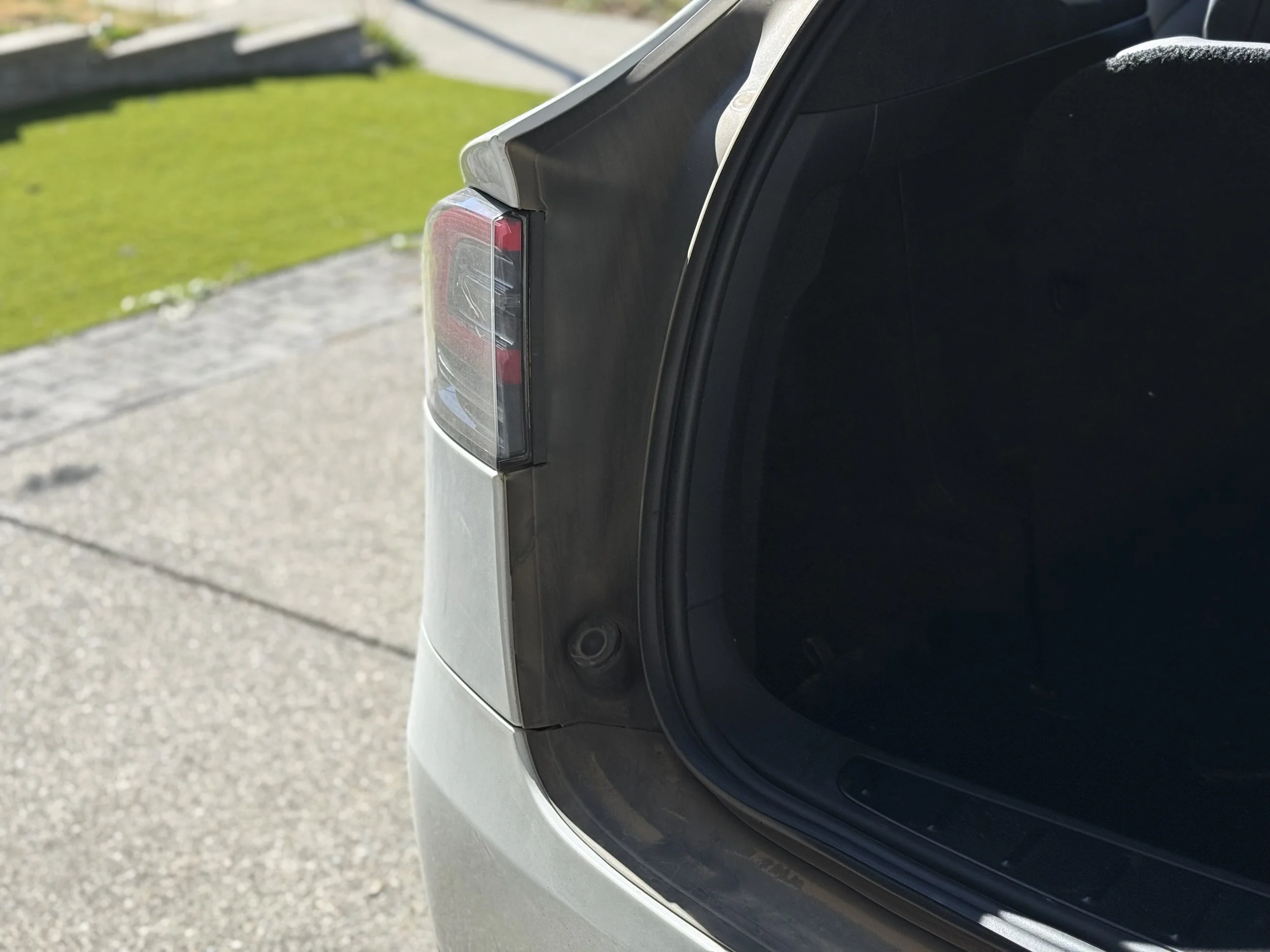Close-up of the rear end of a silver car, showing part of the tail light and the trunk open, parked on a driveway with green grass and a sidewalk in the background.
