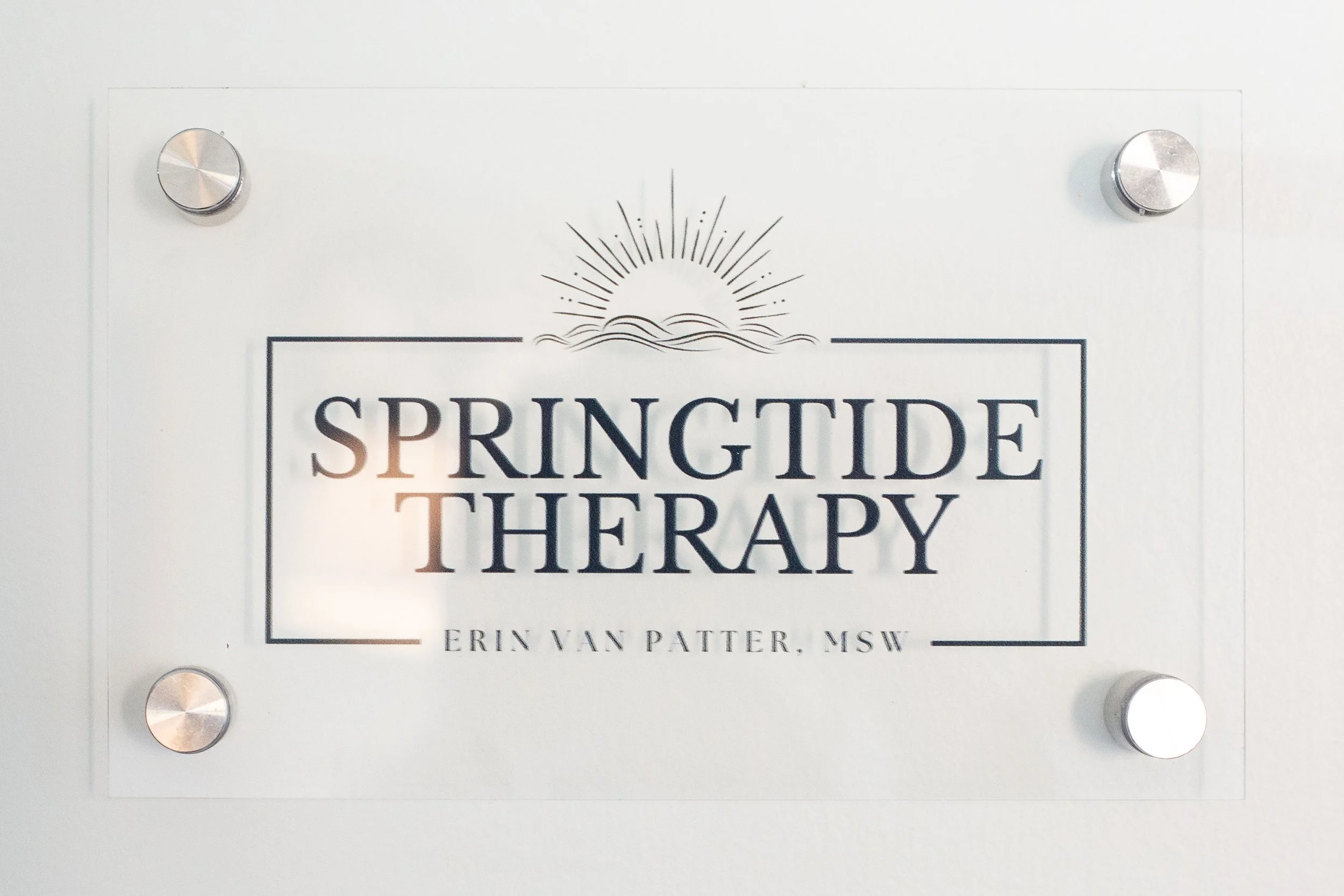 Sign reading 'Springtide Therapy' with the subtitle 'Erin Van Patter, MSW' and a simple sun illustration at the top, mounted with four metallic bolts.