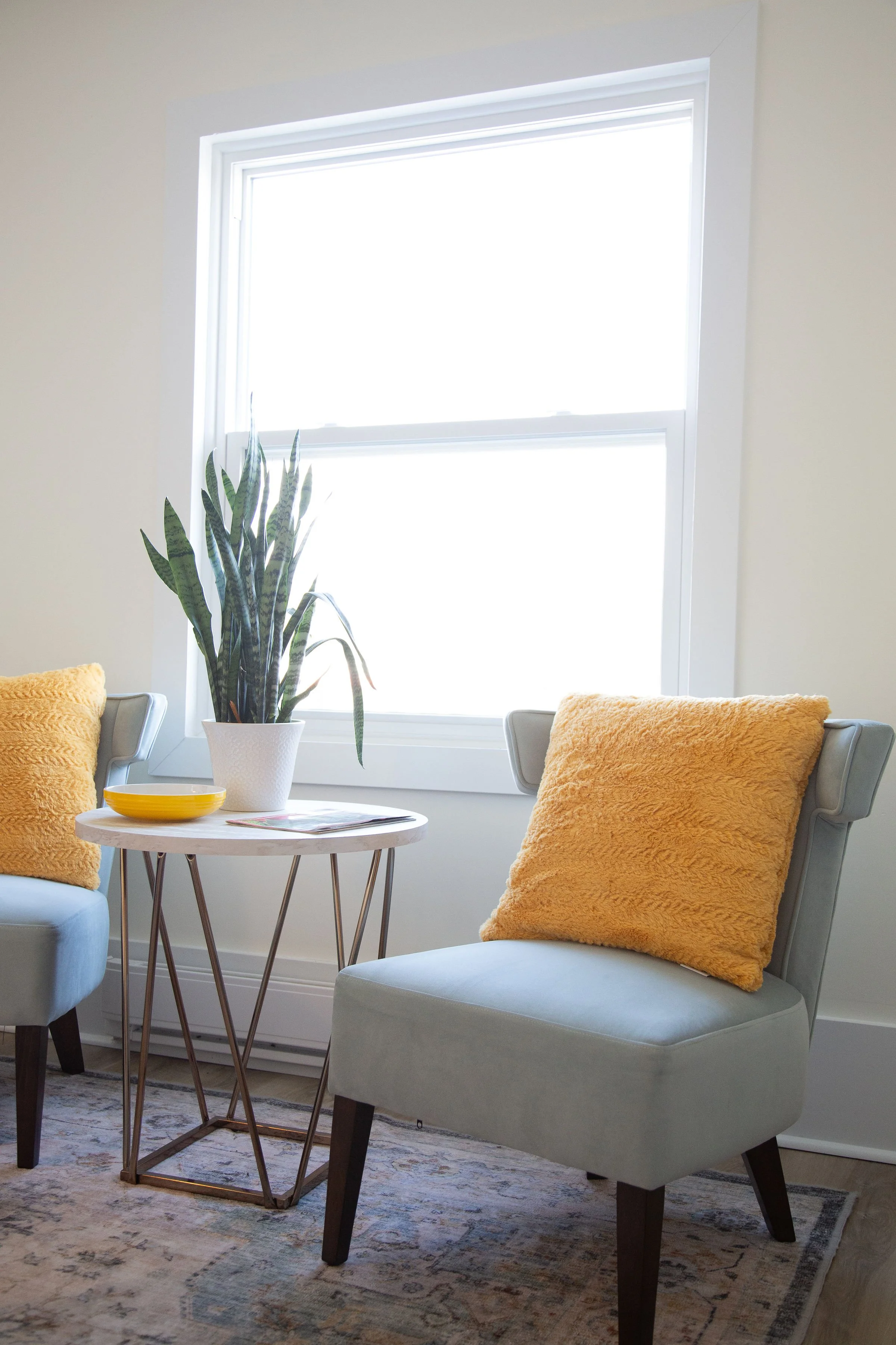 Living room with a window, a light gray armchair with yellow pillows, a small wooden side table with a metal base, a potted snake plant, a bowl, and a magazine, a rug with a traditional pattern, and light-colored walls.
