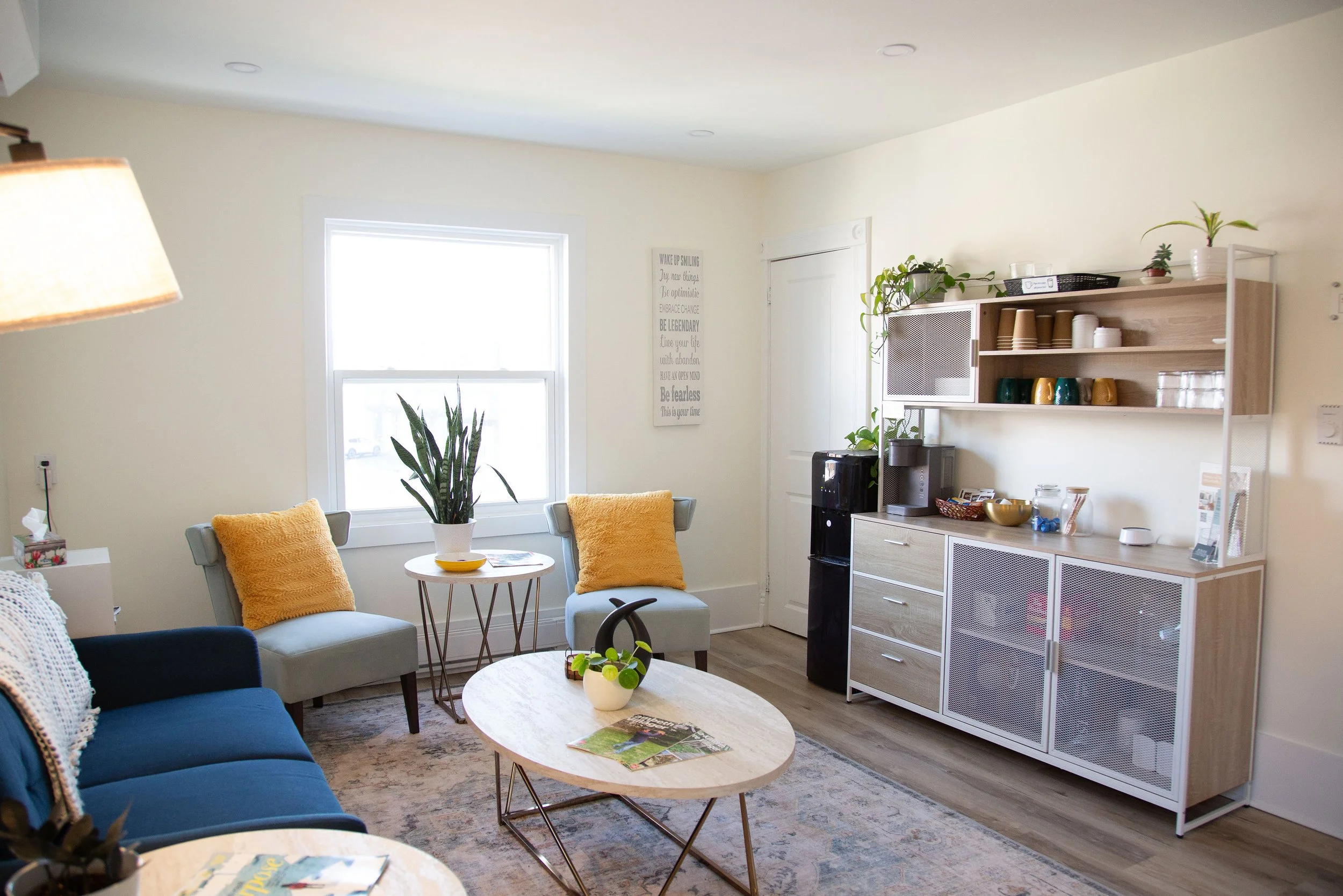 Cozy living room with a blue sofa and yellow pillows, a round coffee table, potted plants, and a wall shelf with cups and decor, sunlight coming through a window.