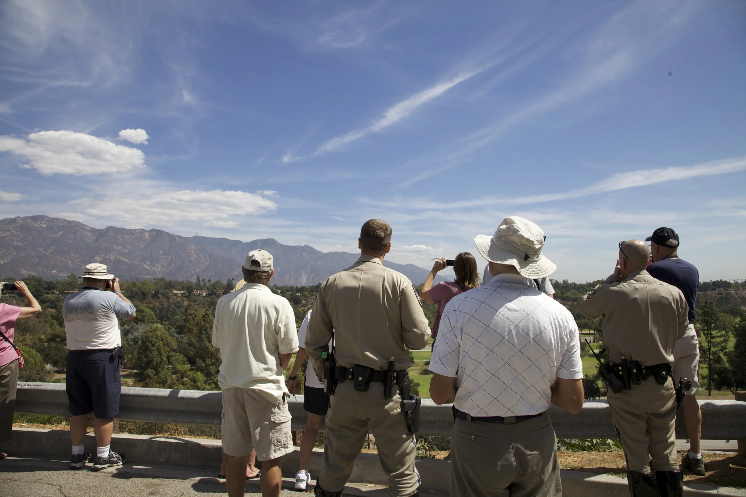 Group of people, including some in uniforms, standing on a roadside overlook, looking at mountains and landscape under a blue sky.