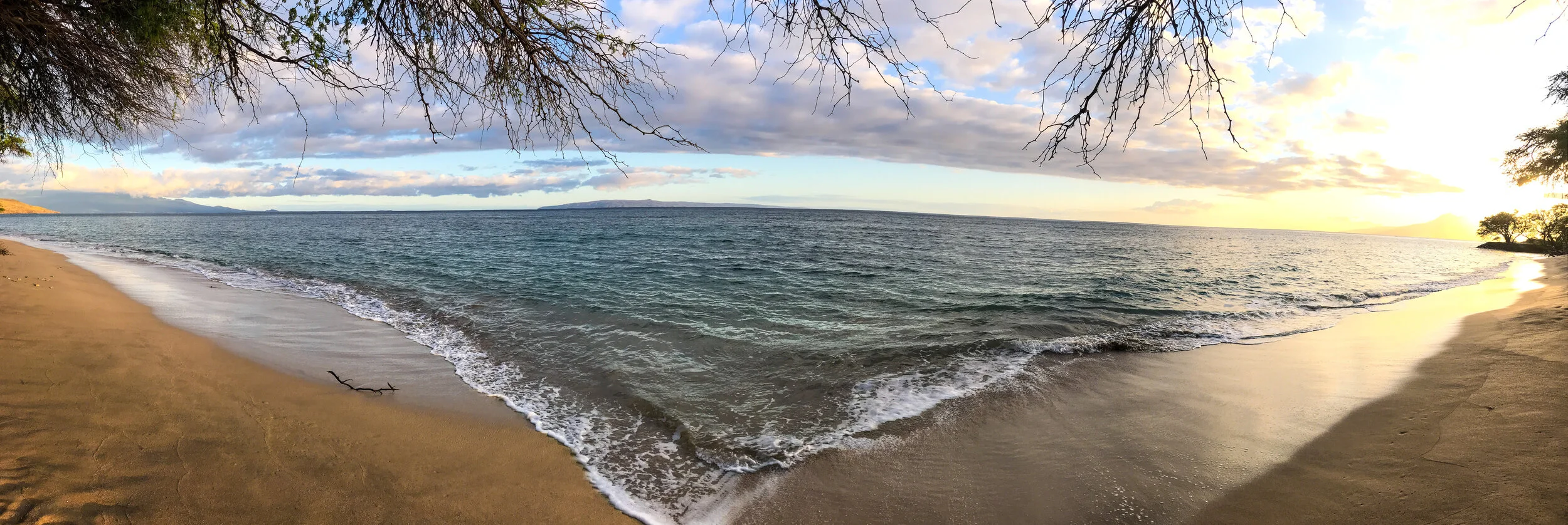 A scenic view of a beach with gentle waves, sandy shore, and trees with bare branches under a partly cloudy sky, with the sun setting on the horizon.