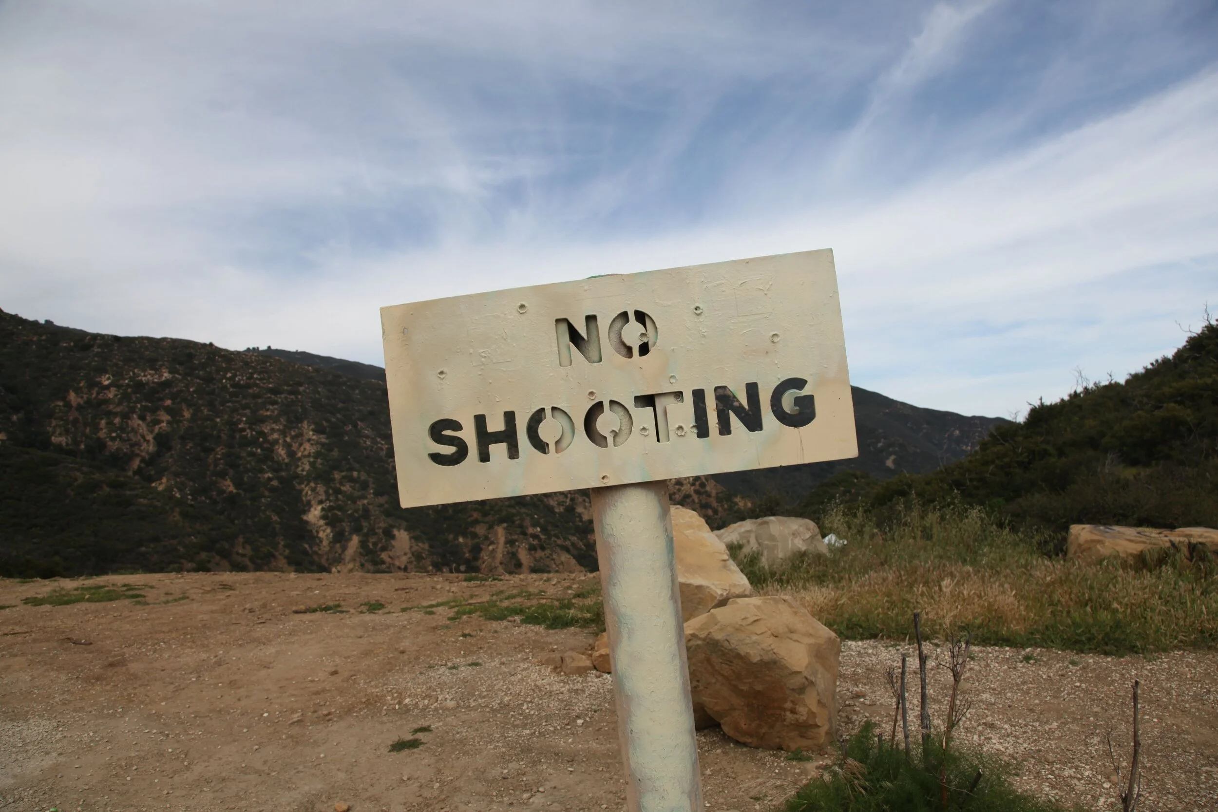 A weathered sign on a post that reads "No Shooting" in a rugged outdoor landscape with hills, rocks, and a partly cloudy sky.