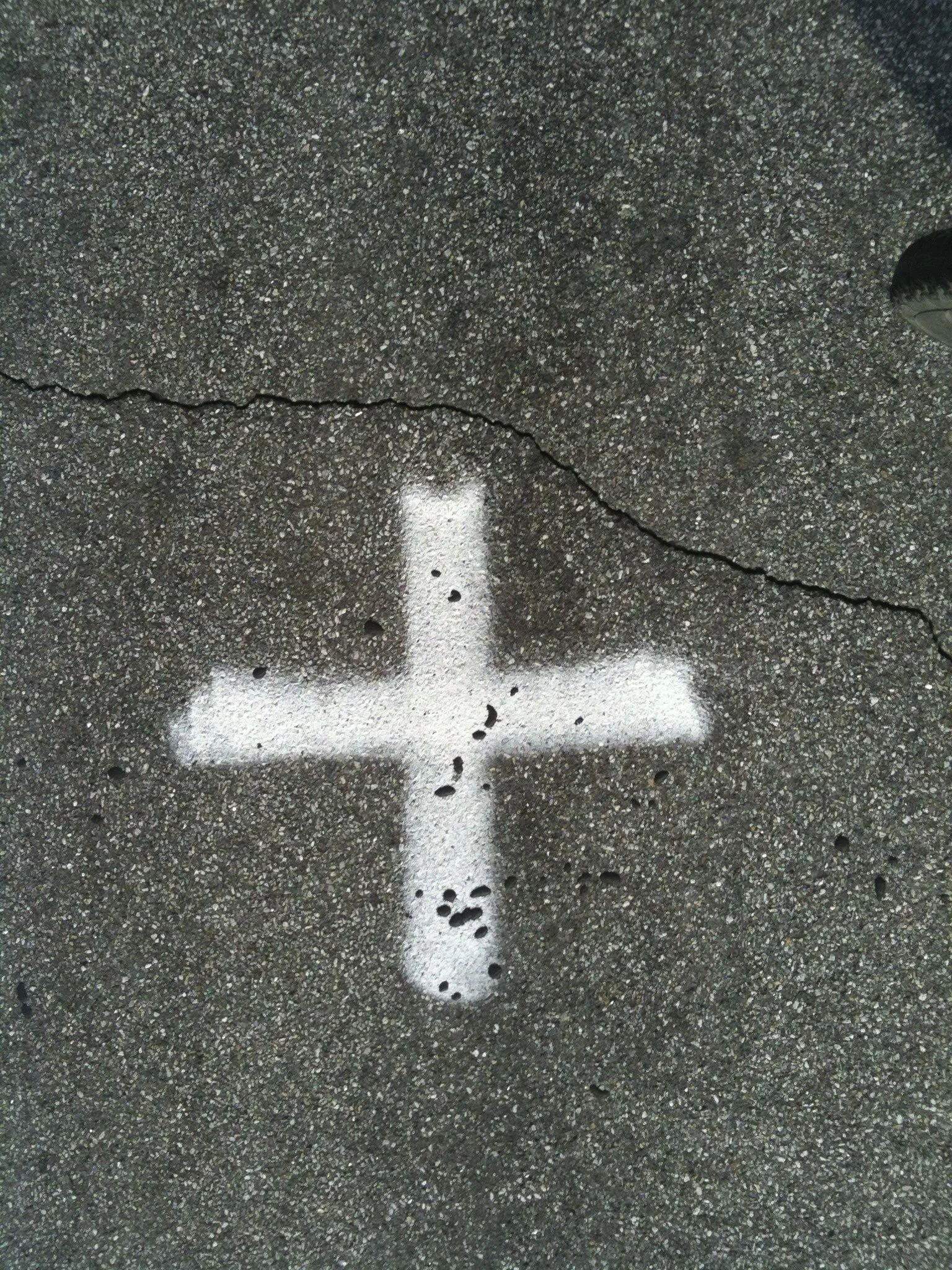 White spray-painted plus sign on asphalt pavement with small holes and a visible crack.