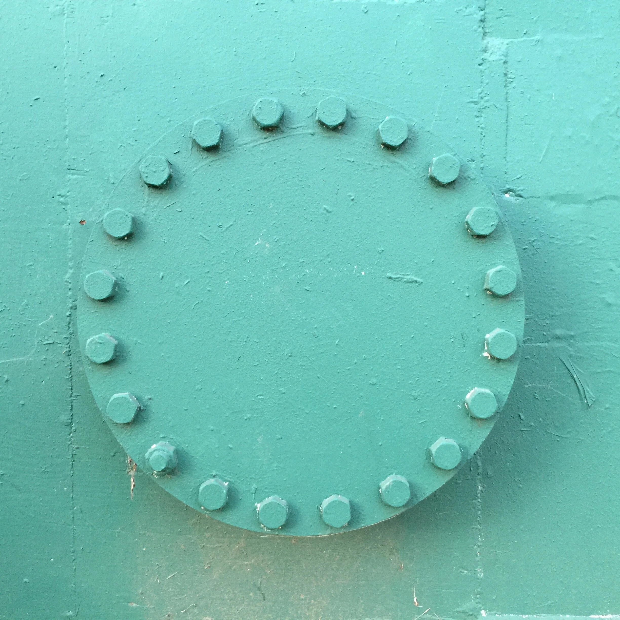 A close-up view of a round, teal-colored metal cover with evenly spaced bolts around its perimeter, on a matching teal background.