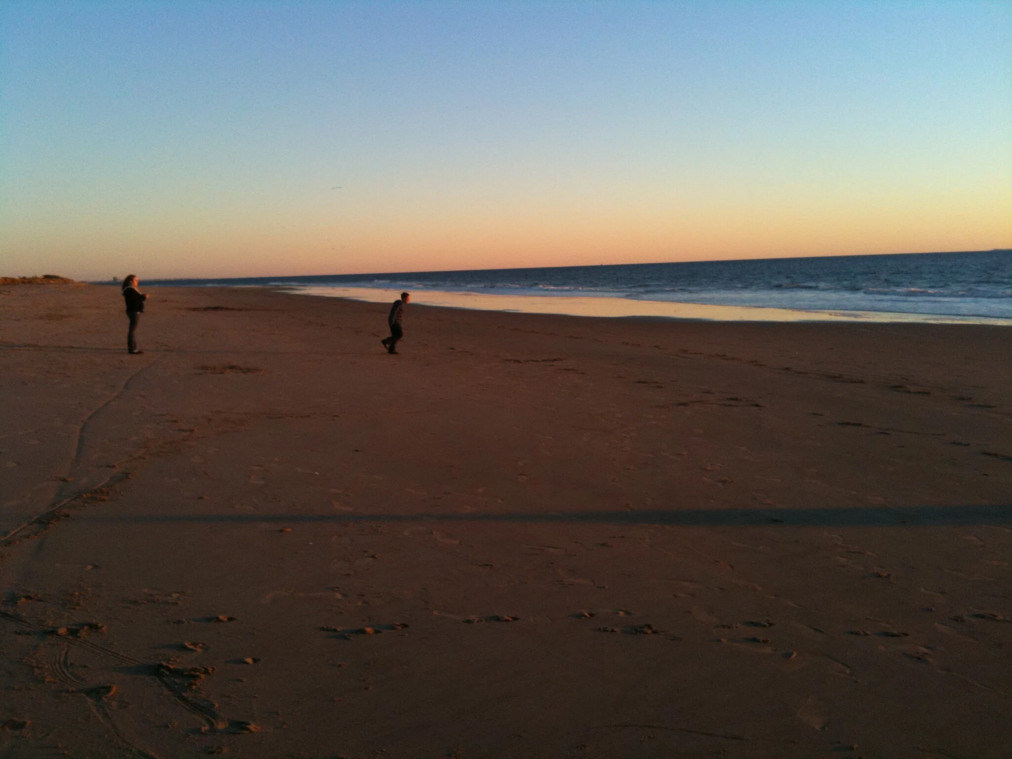 People walking on a sandy beach at sunset, with the ocean waves approaching the shore and the sky transitioning from blue to pinkish hues.