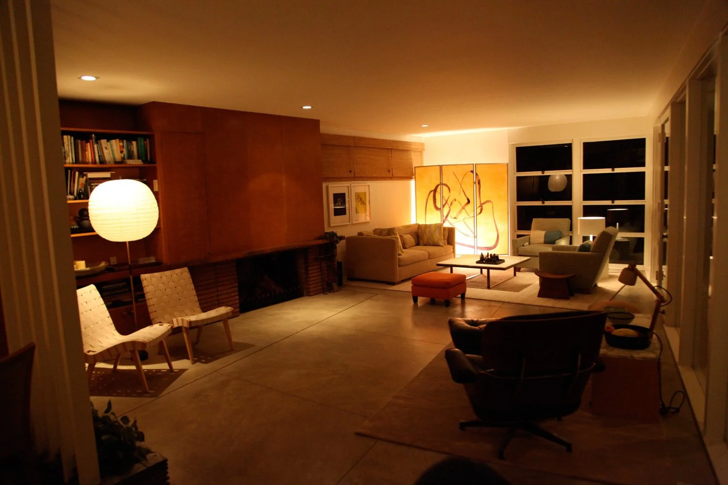 Living room at night featuring a fireplace, beige sofa, armchairs, a wooden bookshelf, a floor lamp, and artwork with warm lighting.
