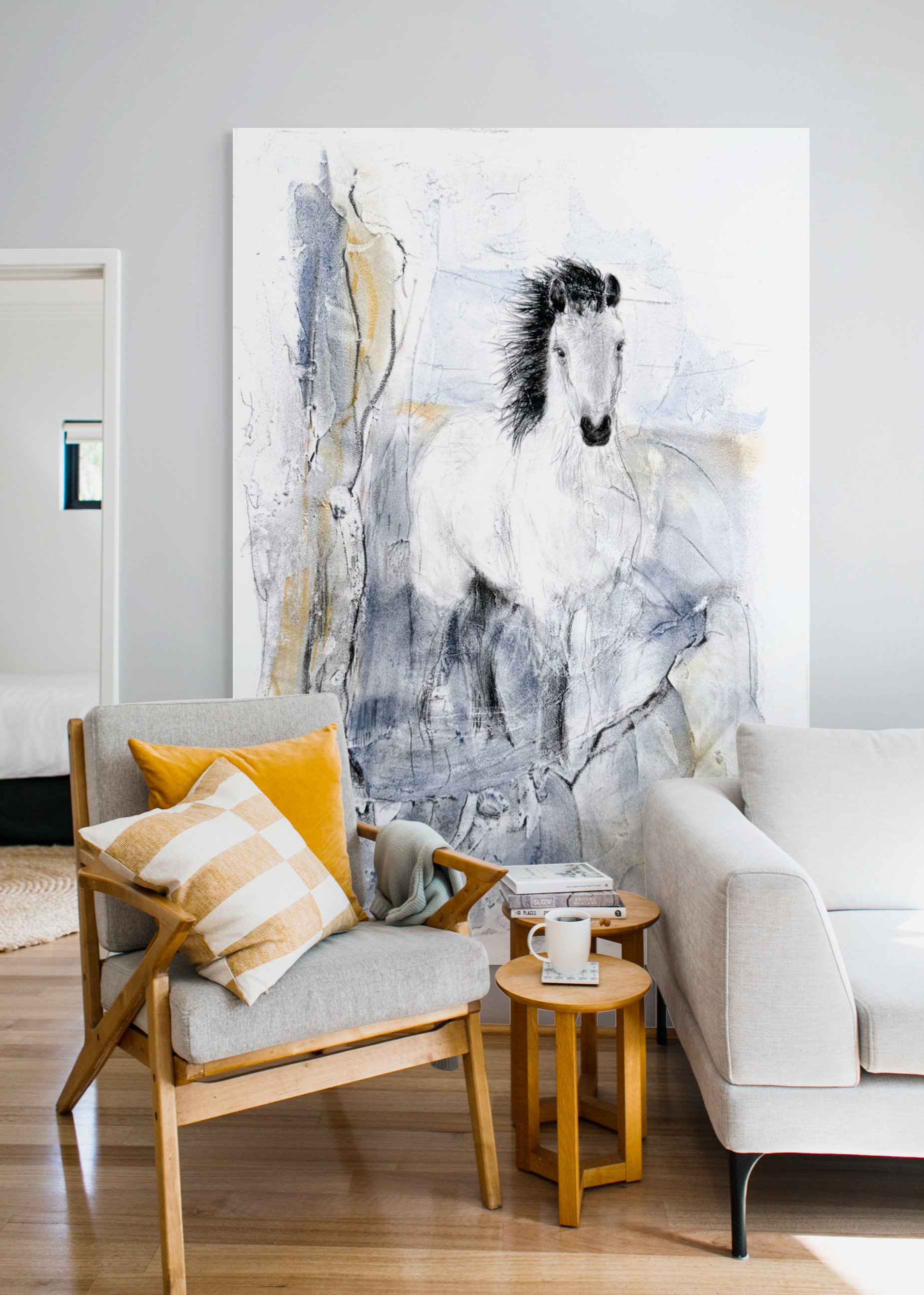 Living room with a large wall art featuring a horse and a tree. There is a gray armchair with a yellow pillow and a striped pillow, and small wooden tables next to it. A white couch is partially visible alongside a coffee mug and books on the tables.