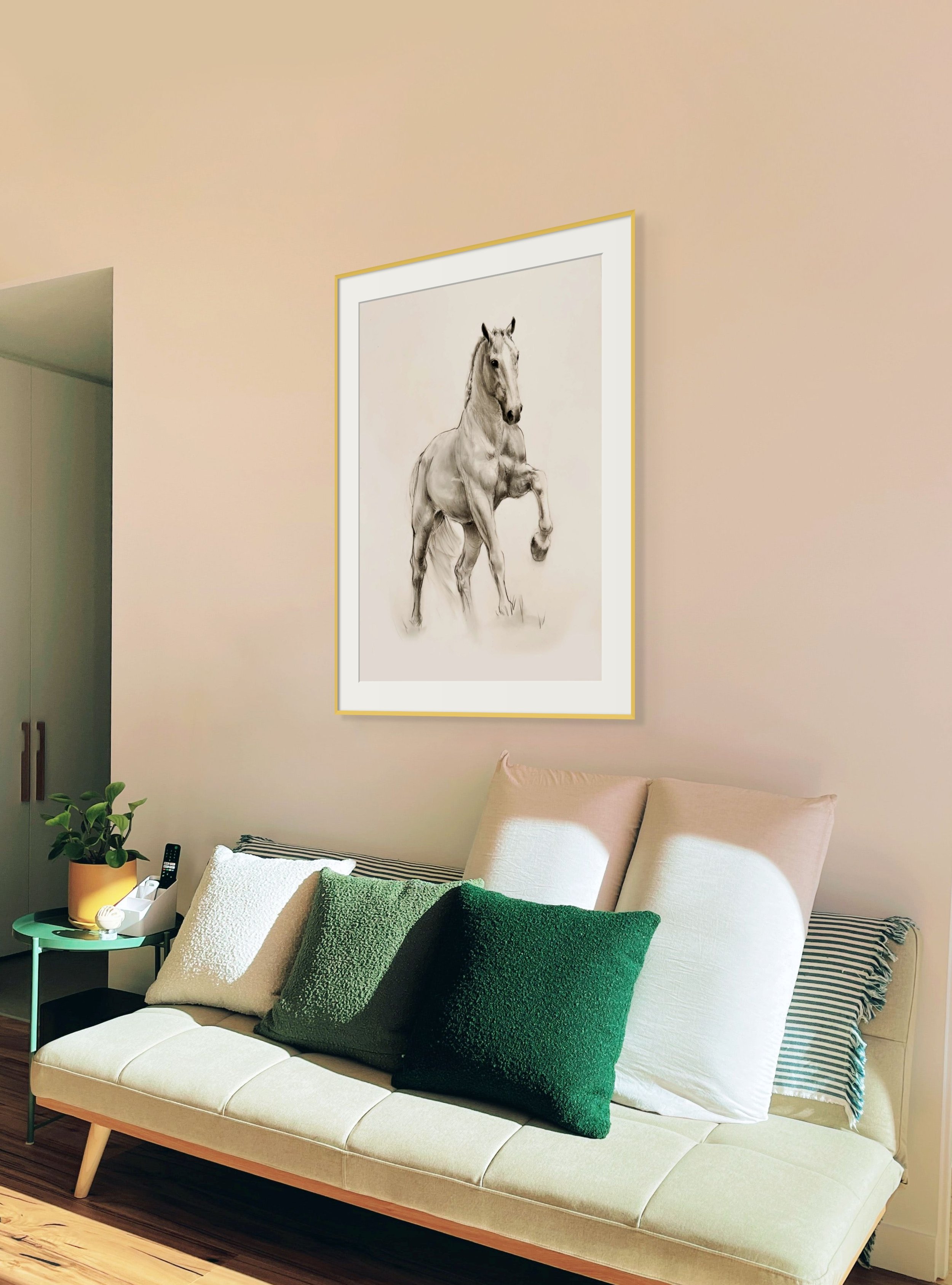 Living room with a beige sofa adorned with white and dark green cushions, a small green side table with a potted plant and remote control, and a framed black-and-white horse drawing on the wall.