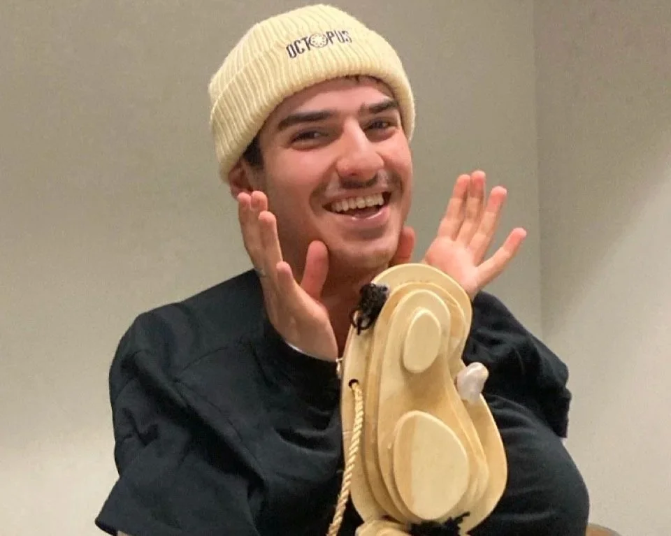 A young man smiling with one hand near his face, wearing a beige knit beanie and a black jacket, with a wooden phone booth prop in front of him.