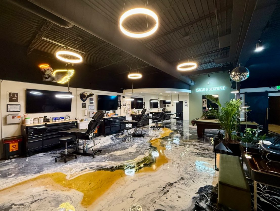 Modern tattoo studio with workstations, large TV screens, circular ceiling lights, a pool table, and a decorative floor resembling a river or path with colorful patterns. Neon sign reading 'SAGE & SERPENT' and mirrors reflecting the interior.