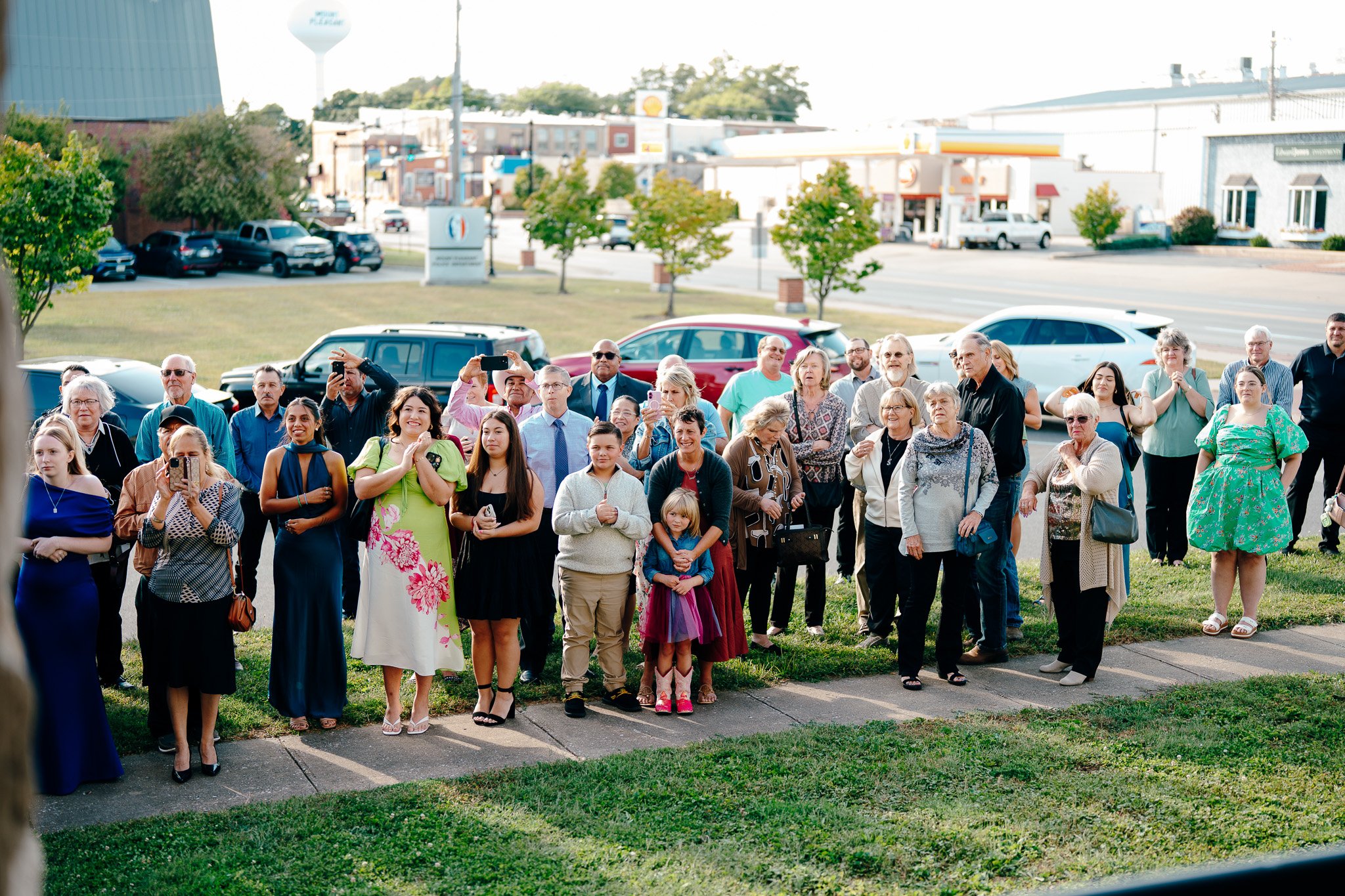 A group of people, including men, women, and children, gathering outdoors on a sidewalk, some taking photos or videos with their phones, in front of parked cars and a city street.