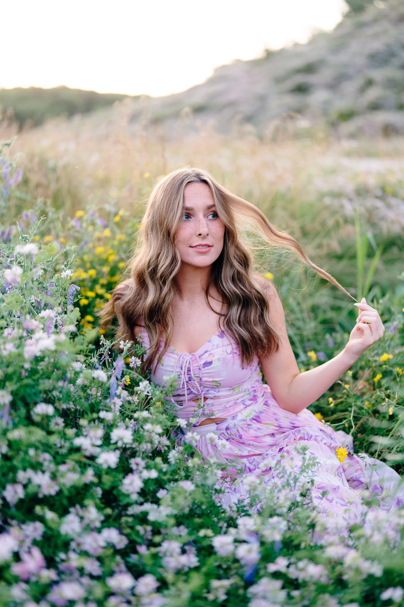 A young woman sitting in a field of wildflowers during sunset, holding a strand of her wavy brown hair.