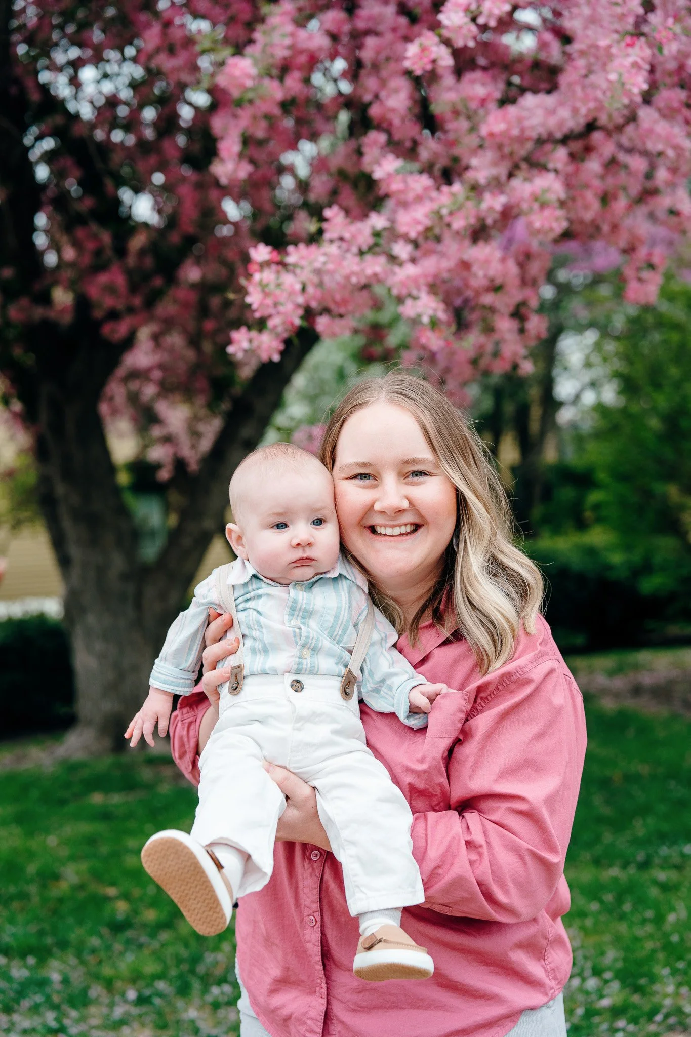 A woman holding a baby in front of a pink blossoming tree outdoors.