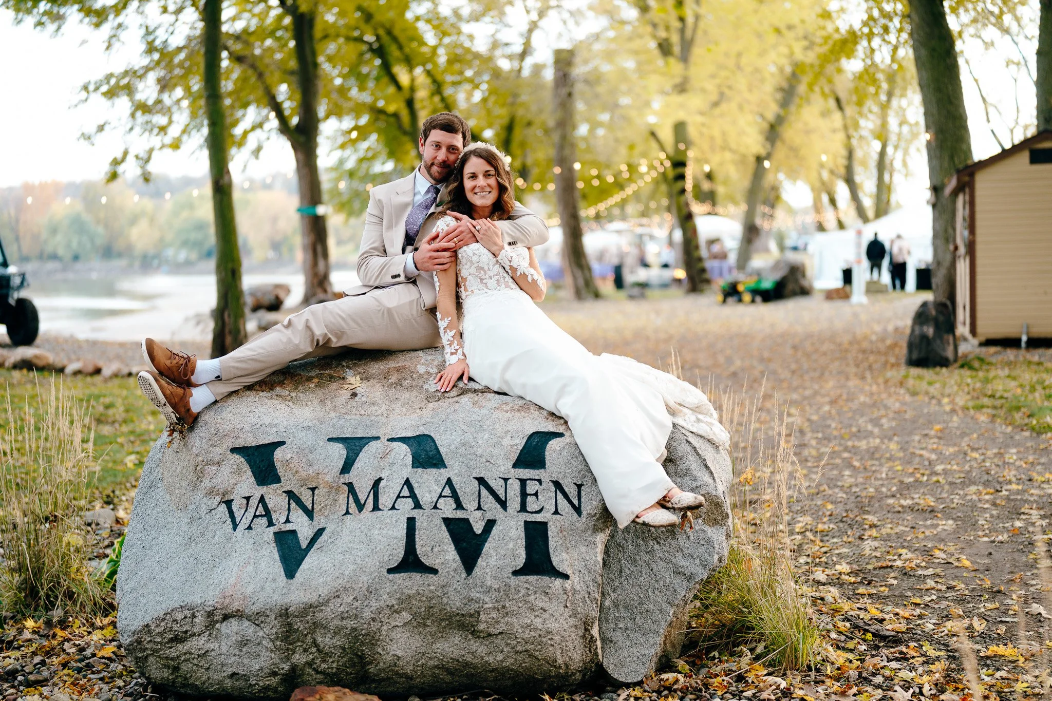 A newlywed couple sitting on a large rock with the words 'VAN MAANEN' and roman numerals engraved, overlooking a park with trees, a lake, and string lights in the background during fall.