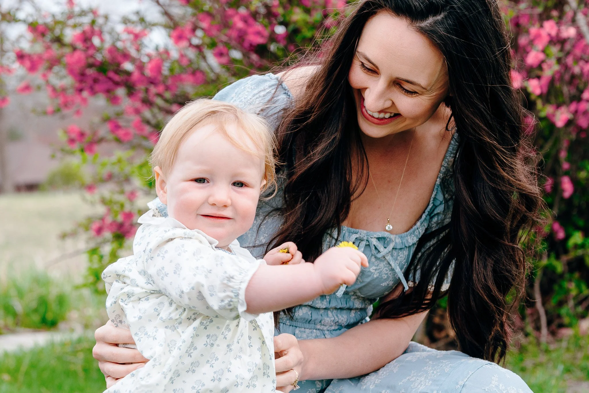 A woman with long dark hair smiling and looking at a young girl with red hair, who is holding a yellow flower. They are outdoors in front of blooming pink flowers.