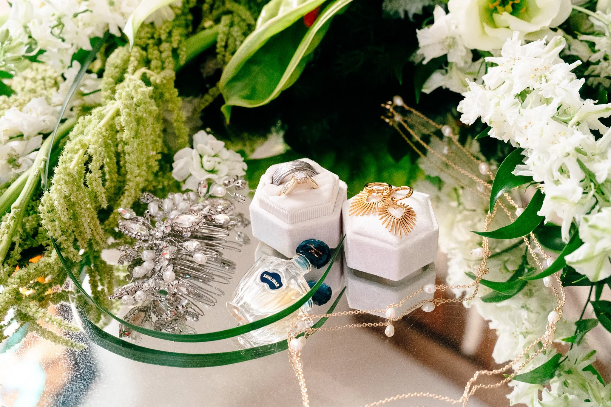 Jewelry display with rings, earrings, necklaces, and a perfume bottle surrounded by white flowers and greenery.