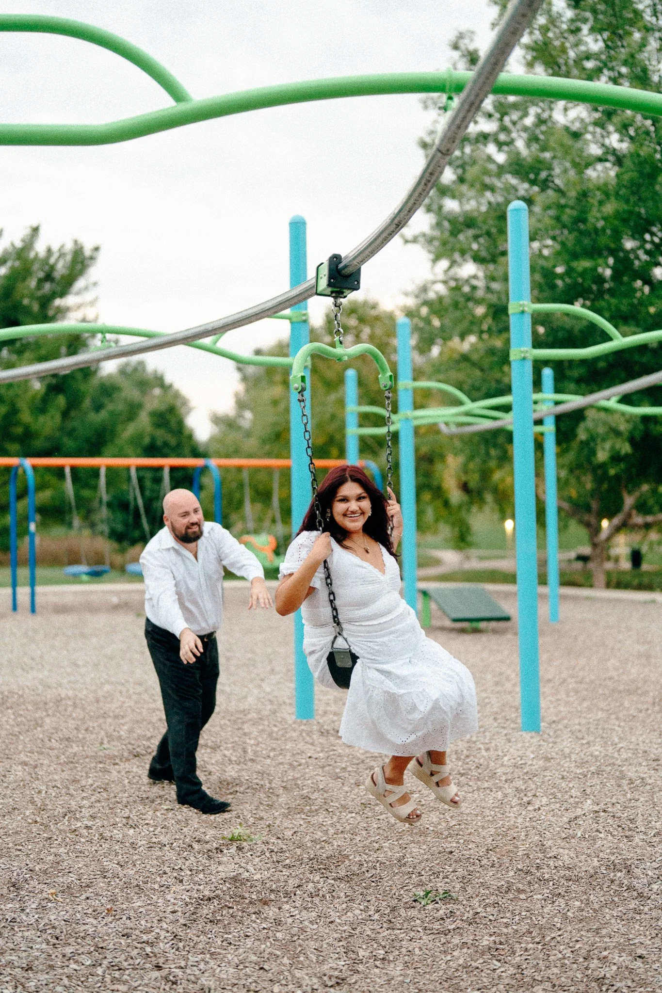 A woman in a white dress smiling on a swing at a playground, while a man in a white shirt and black pants pushes her from behind, both enjoying the moment.