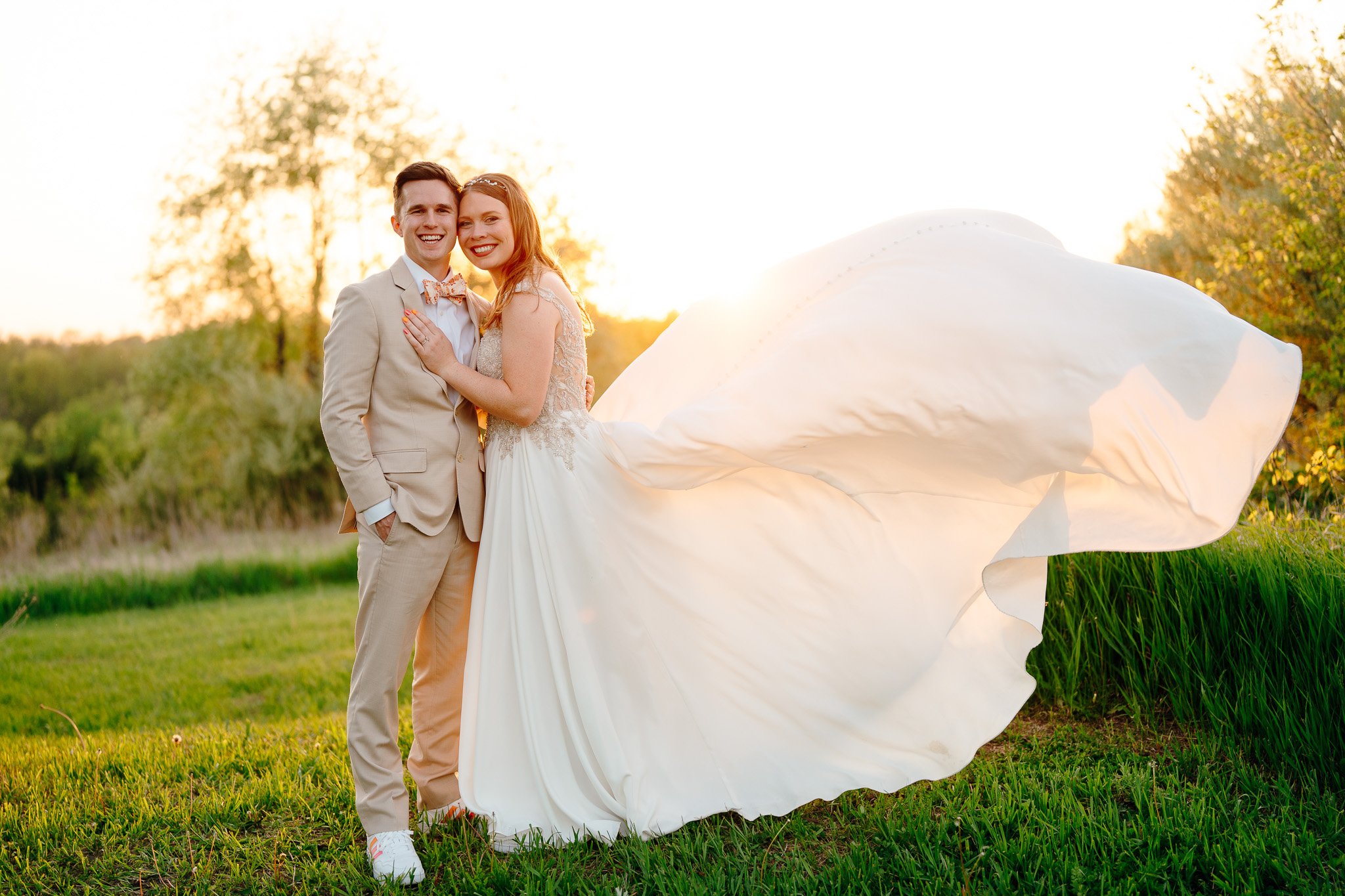 A happy couple in wedding attire standing outdoors in a grassy area during sunset, with the bride wearing a flowing white dress and the groom in a beige suit and bow tie, embracing and smiling at the camera.