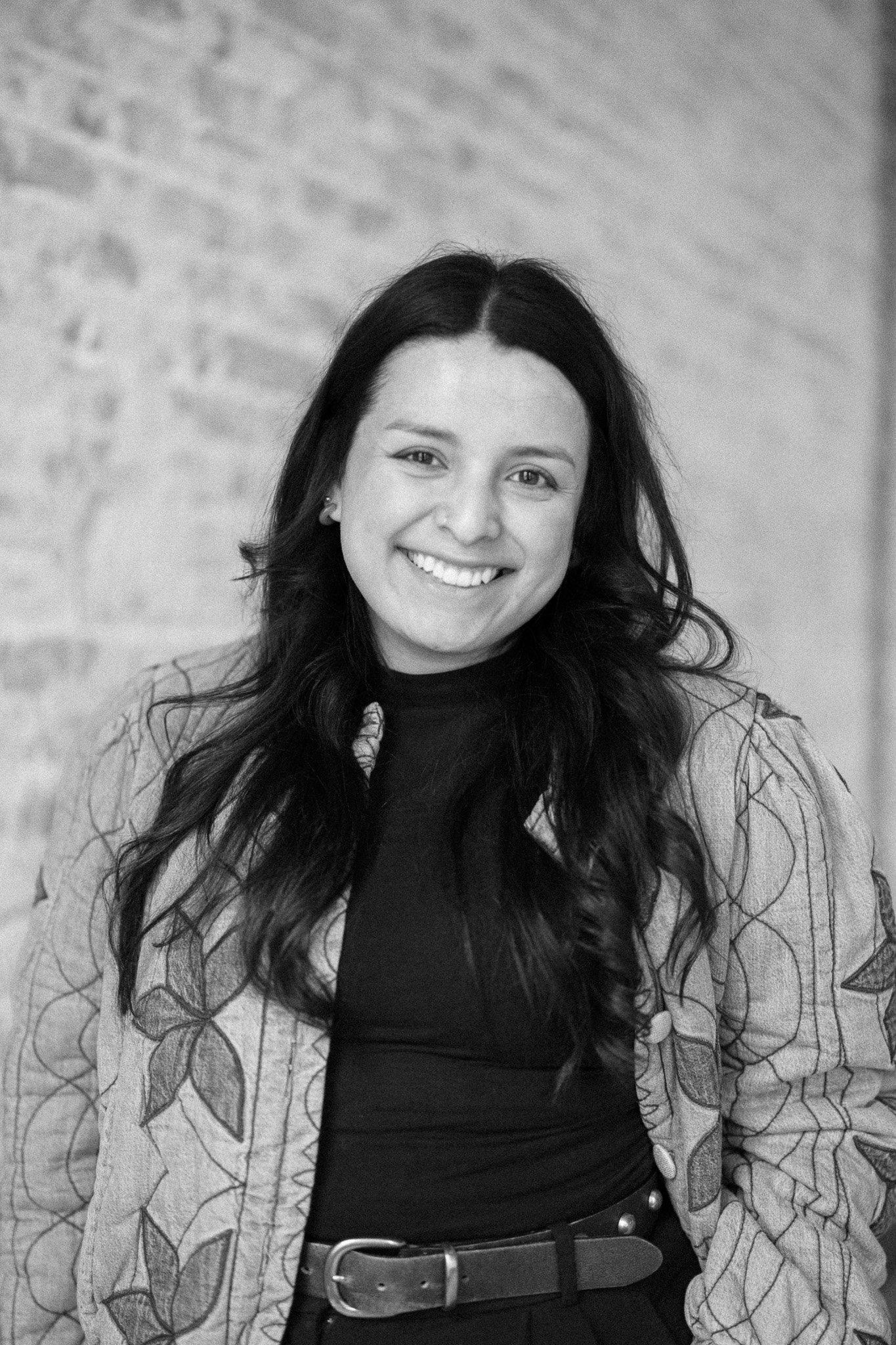 Black and white photo of a smiling woman with long wavy hair, wearing a patterned jacket over a dark top, standing against a brick wall.