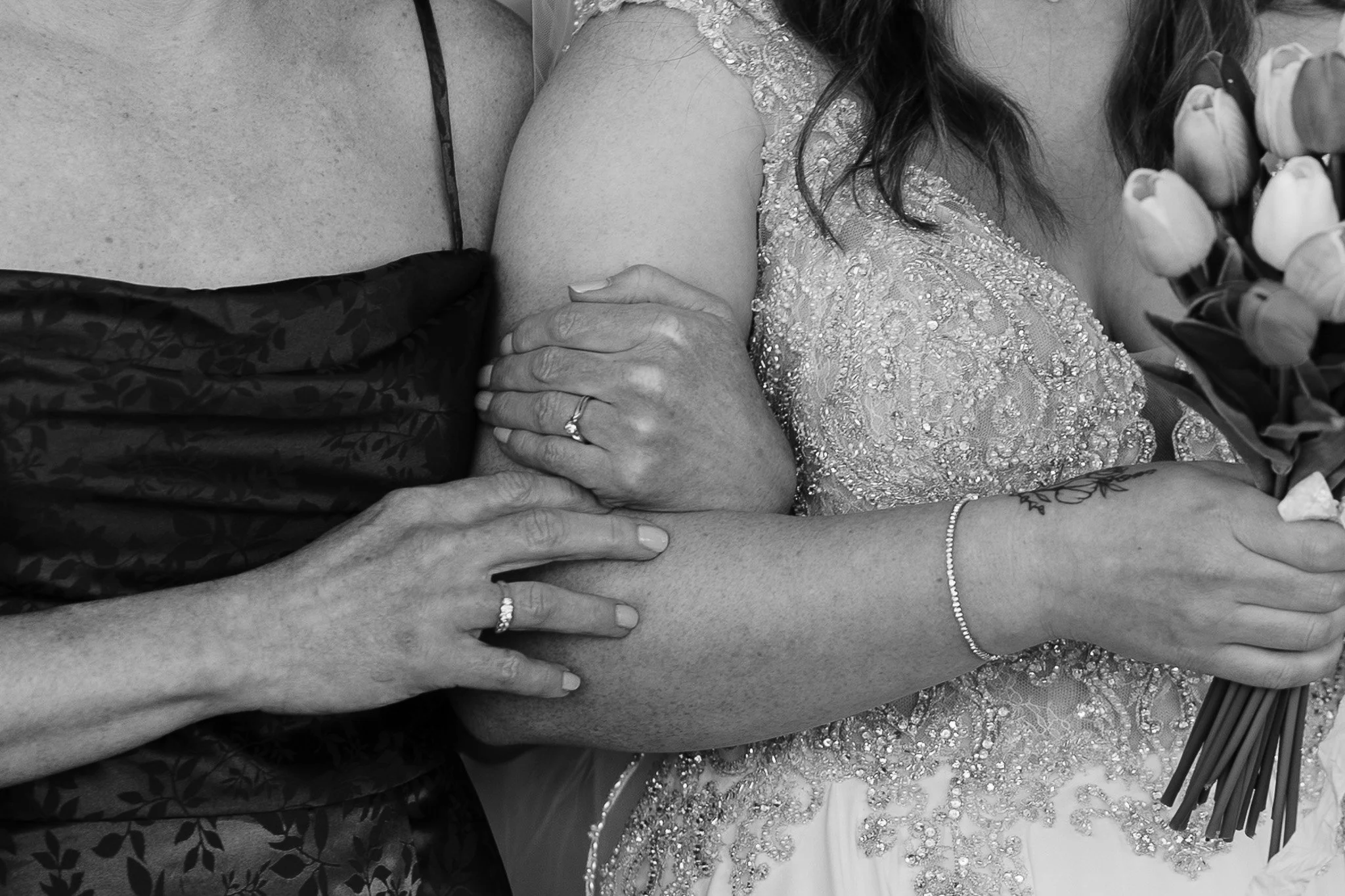 Close-up of two women holding hands and embracing. One woman is wearing a wedding ring, a dress with intricate beadwork, and a bracelet. The other woman's hand rests on her arm, and she wears a ring and a patterned dress. The woman with the beaded dr
