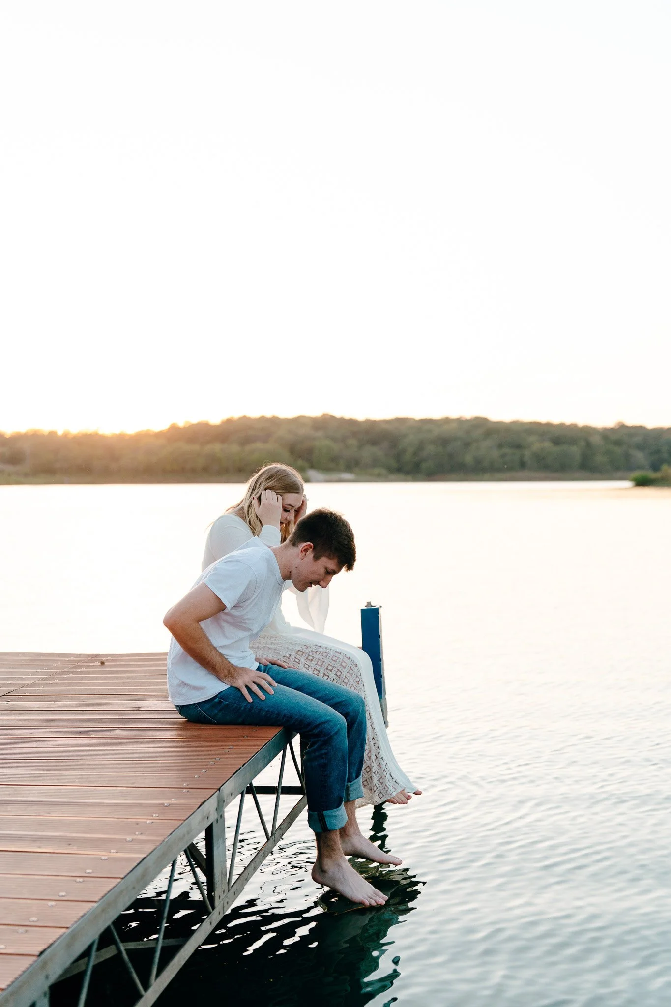 A young couple sitting on a wooden dock by a lake, with their feet in the water, during sunset.