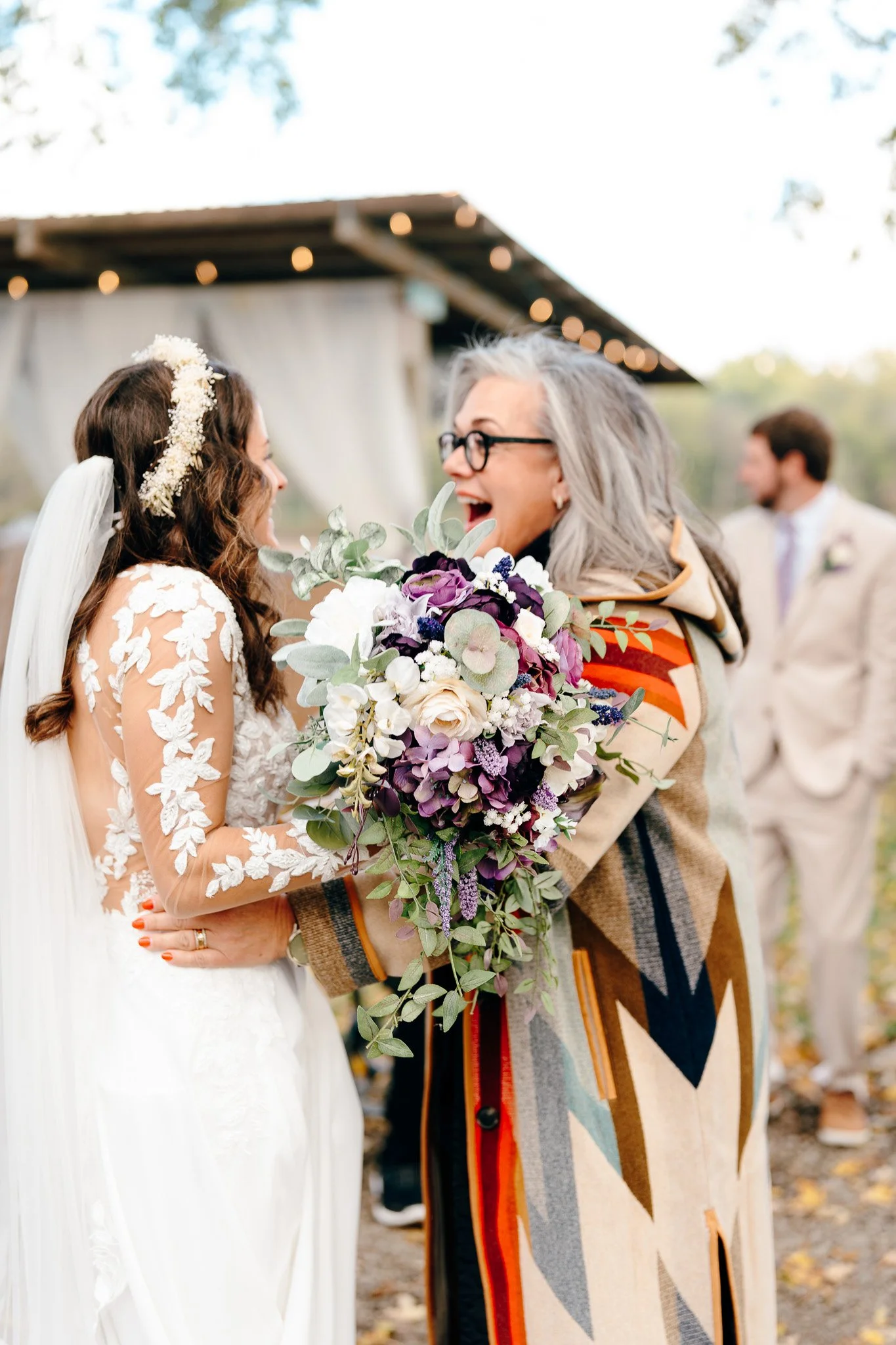 A bride and a woman sharing a joyful moment at a wedding. The woman holds a large bouquet of flowers, and a man and another person are blurred in the background.